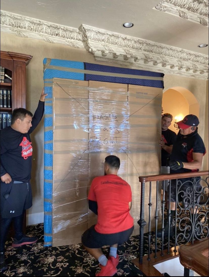 Four workers in uniforms maneuver a large, cardboard-wrapped item near a wooden staircase inside a room.