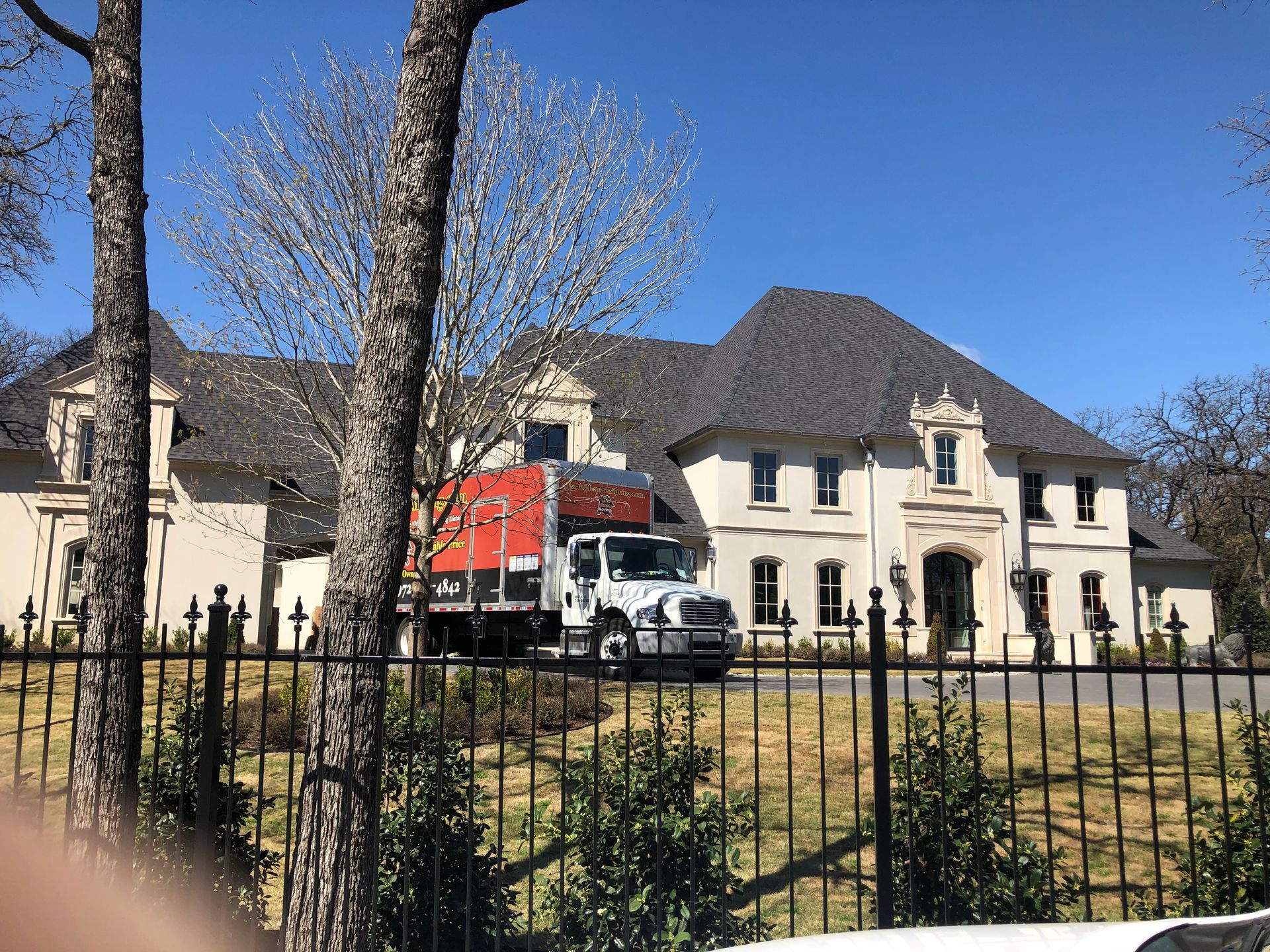 A white box truck parked in the driveway of a large, multi-story Mediterranean-style home behind a metal fence.