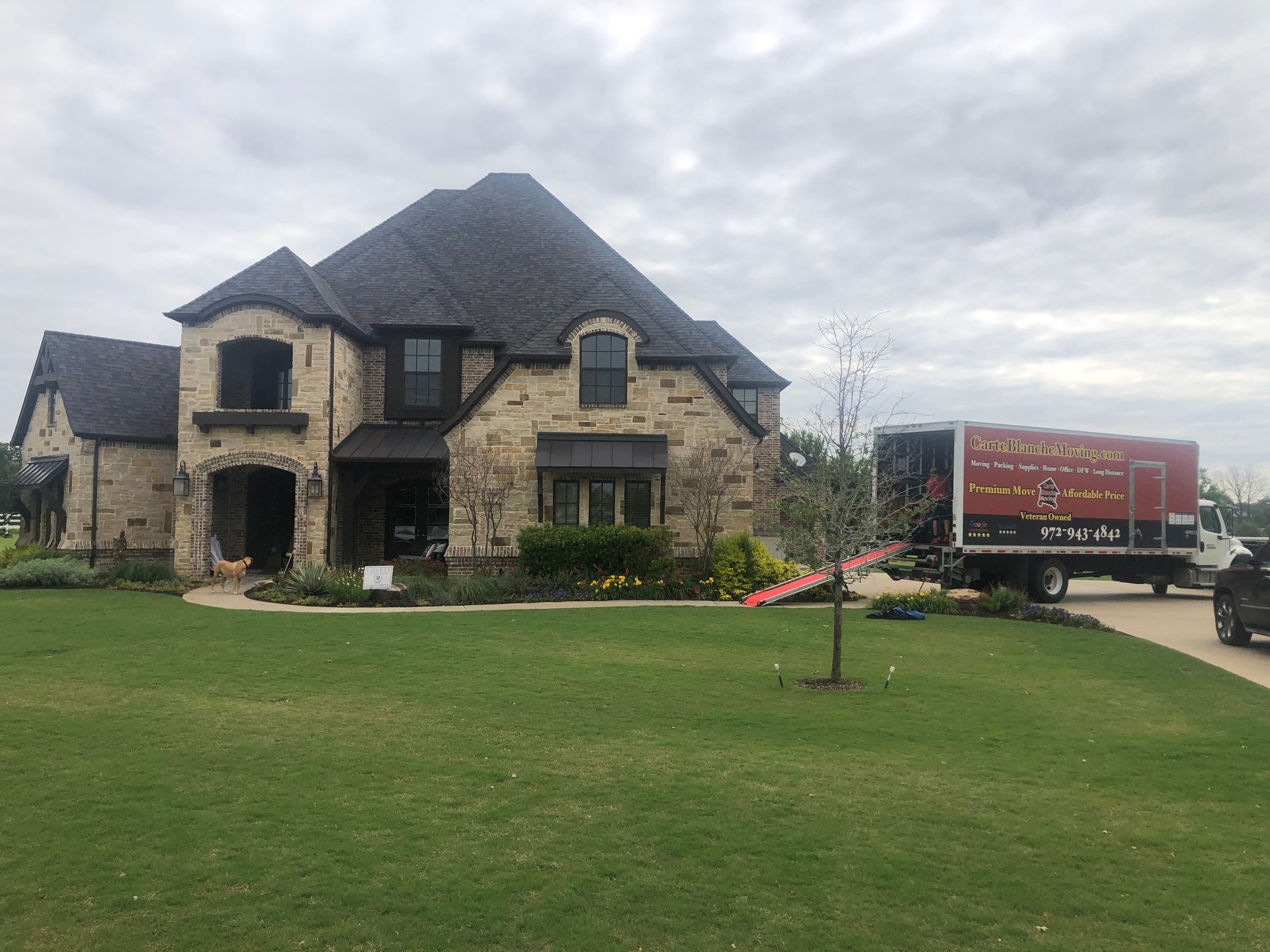 A large stone home with a dark shingled roof under a cloudy sky, with a moving truck parked in the driveway.