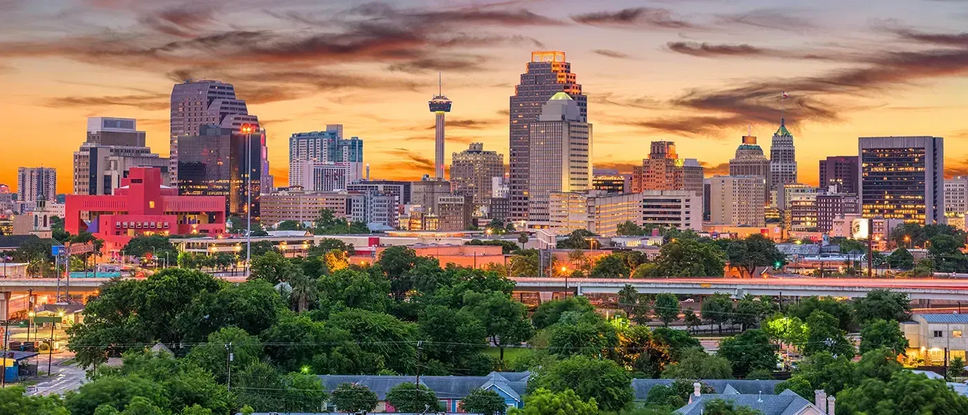 San Antonio, Texas skyline at sunset with the Tower of the Americas visible among buildings and lush green foreground.