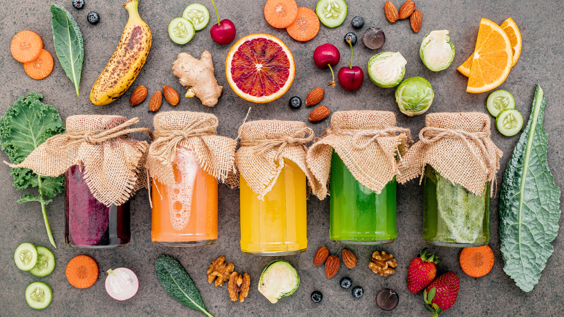 There are many different types of juices in jars surrounded by fruits and vegetables.