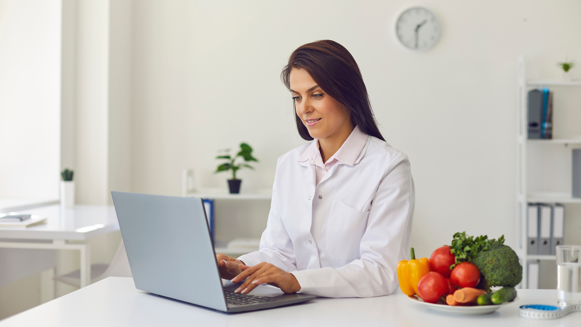 A nutritionist in a white coat working on a laptop with vegetables on the desk.