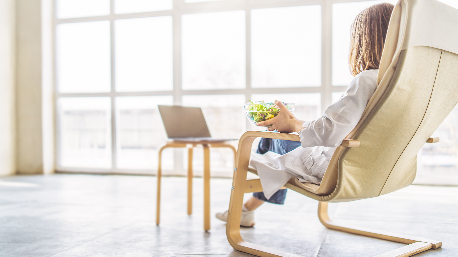 Woman in chair by window, laptop and salad bowl on small table. Sunlight.