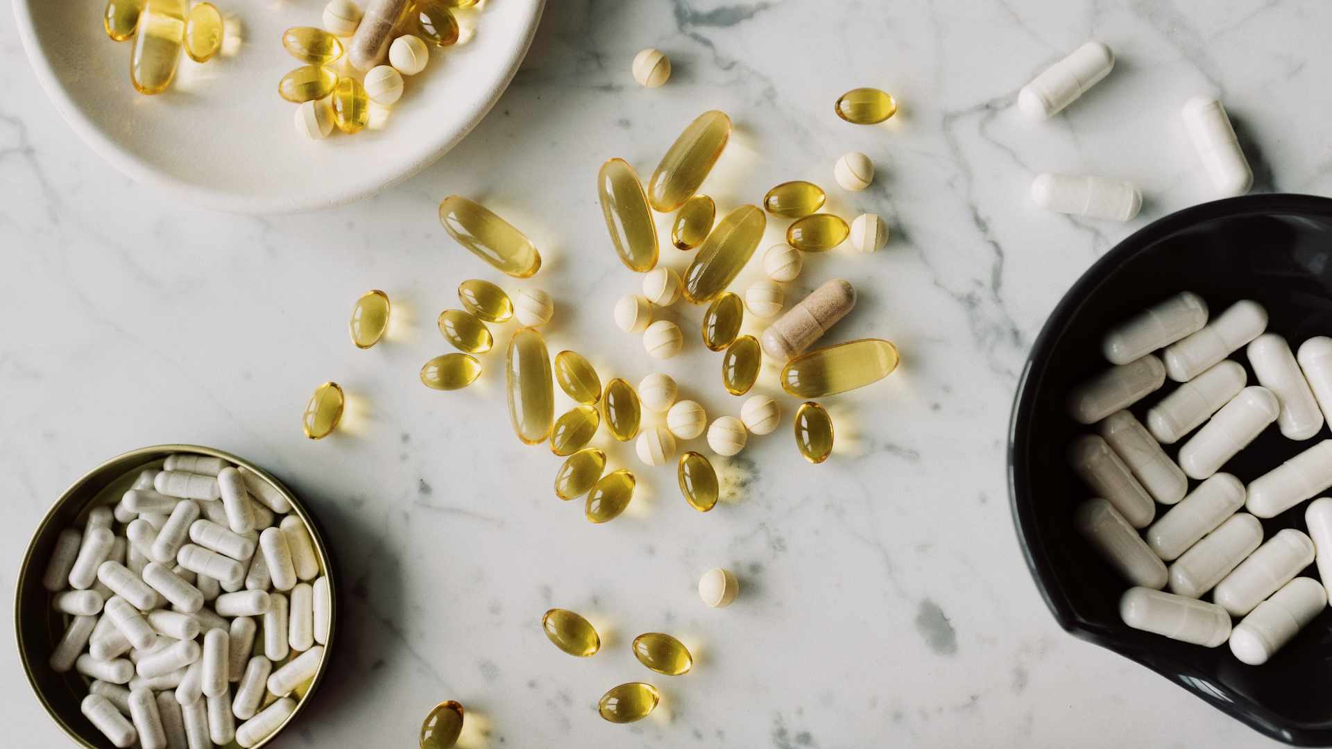 Assorted pills and capsules on a marble surface in small bowls and scattered around, various colors.