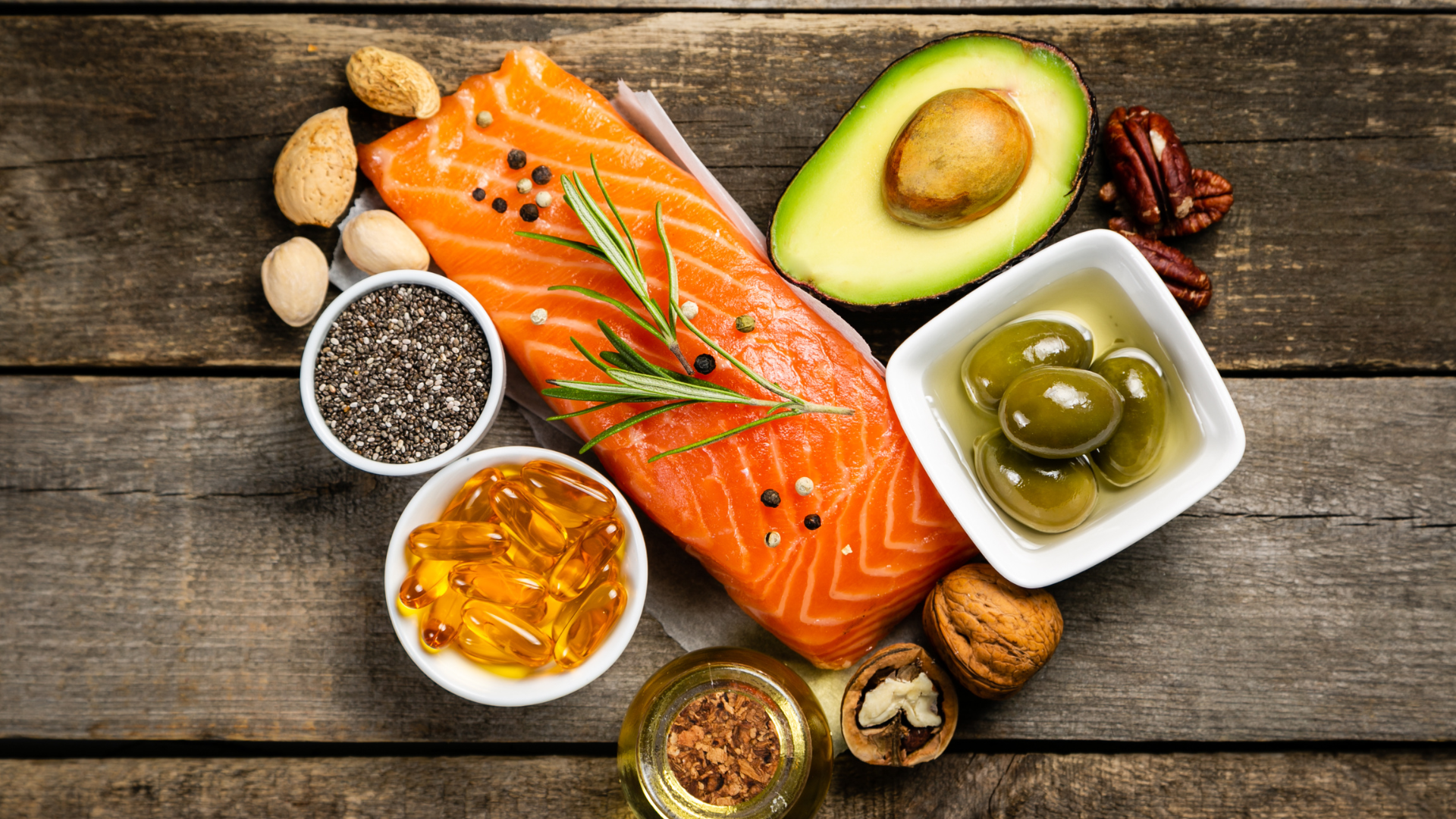 A wooden table topped with salmon , avocado , olives , chia seeds and other healthy foods.