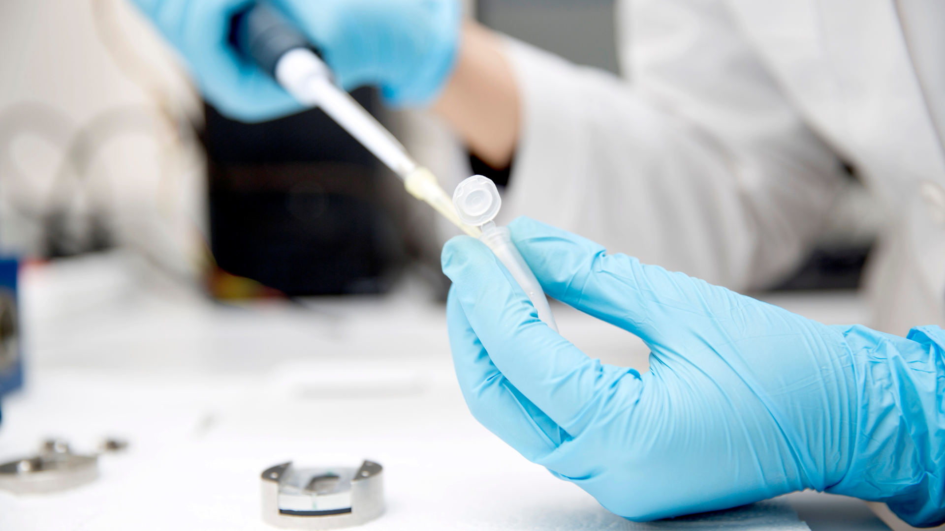 Person in blue gloves using a pipette to dispense liquid into a test tube in a lab setting.
