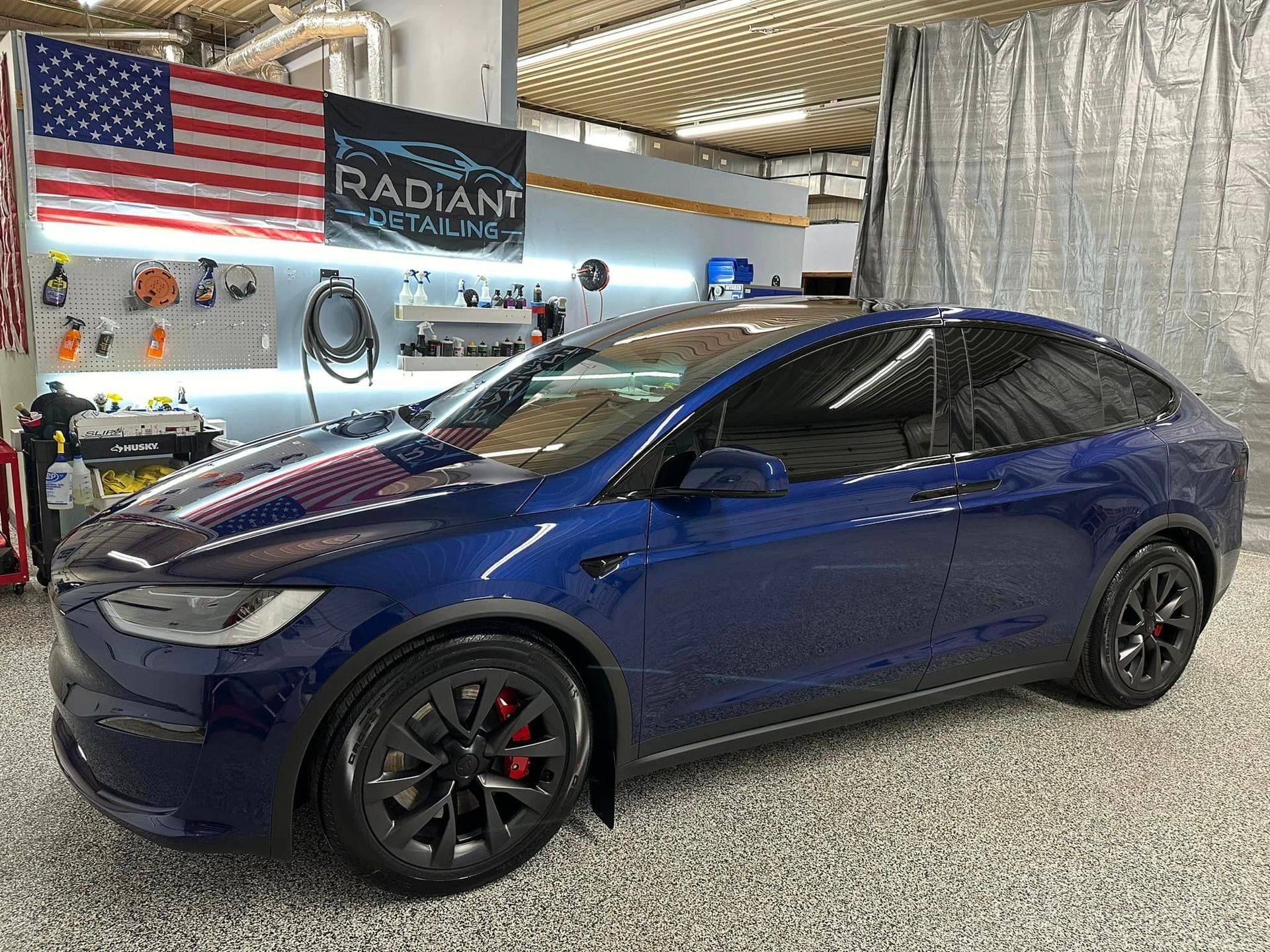 A blue tesla model x is parked in a garage next to an american flag.