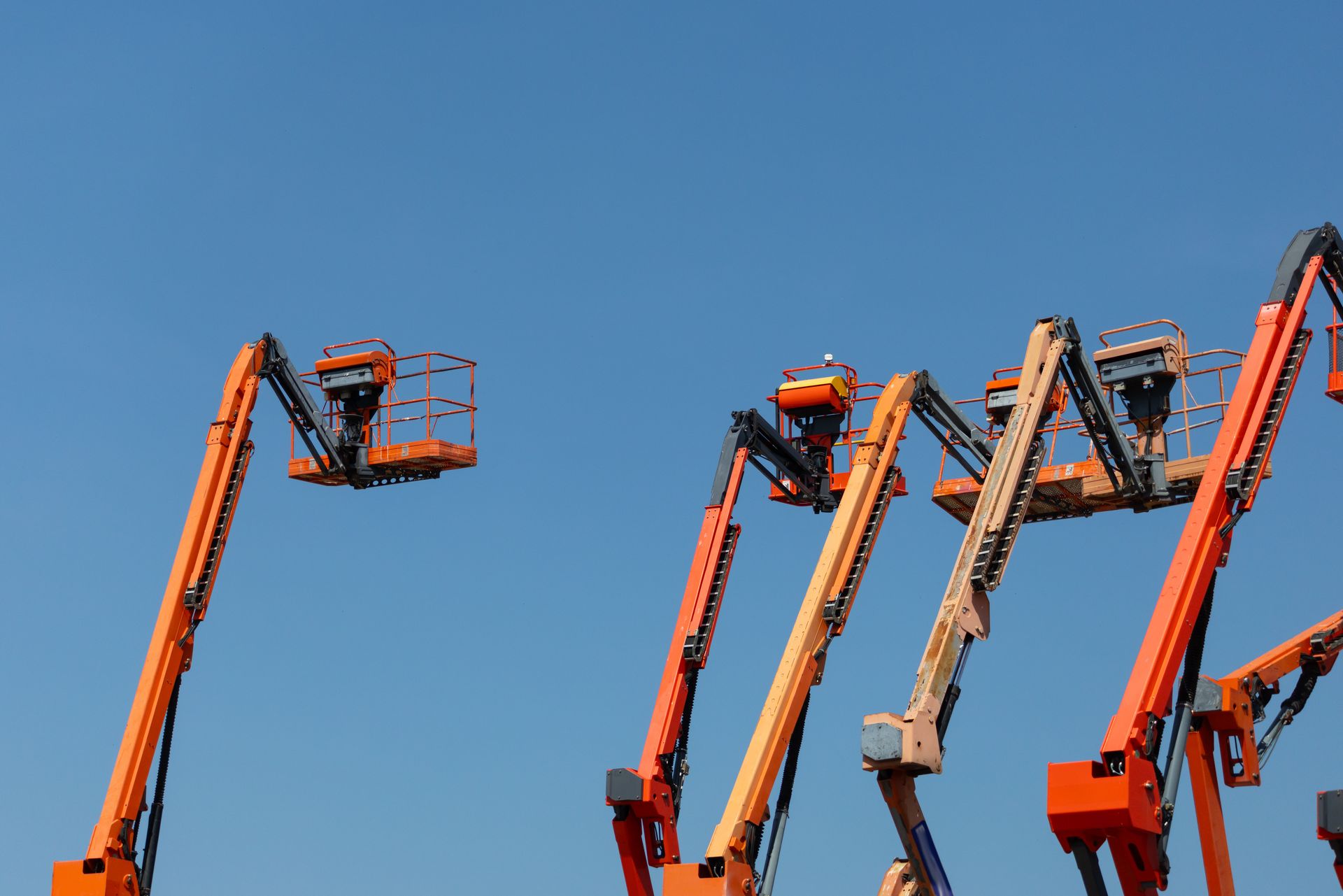 Orange hydraulic boom lifts with raised platforms against a clear blue sky.