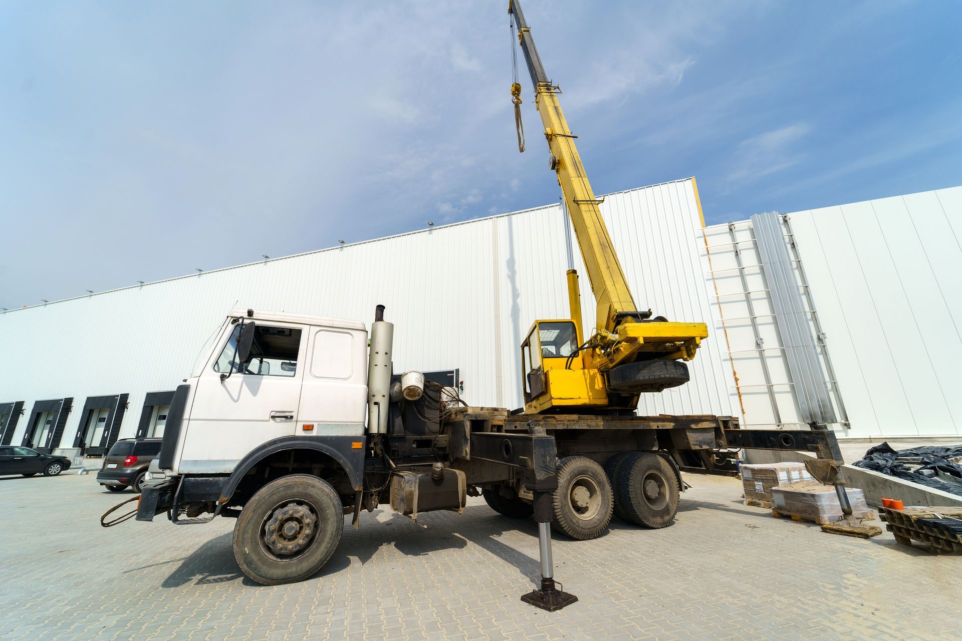 Crane rental truck parked near large warehouse building under clear blue sky on industrial site. Crane rental truck parked near large warehouse building under clear blue sky on industrial site.
