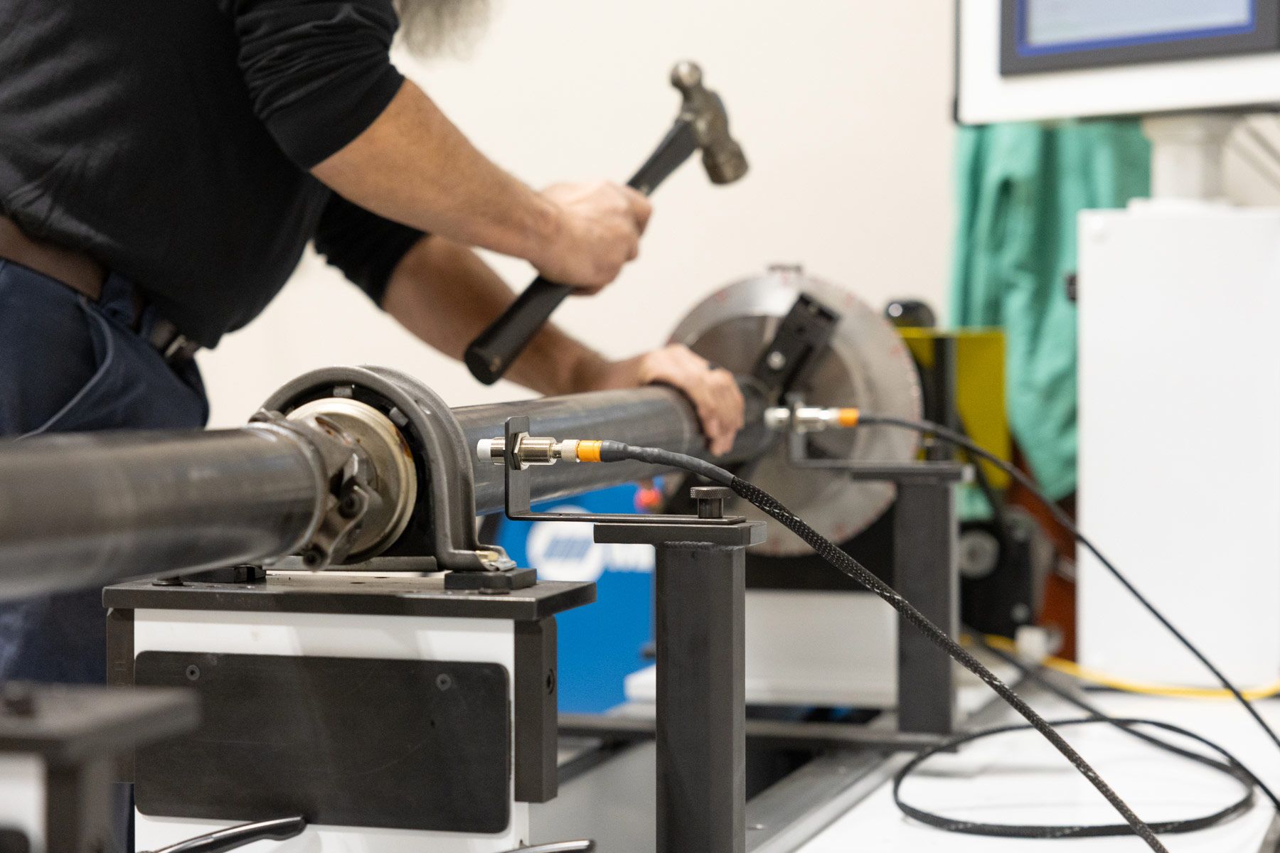 A mechanic works on metal with a yellow tool in a workshop. He wears glasses and a shirt with a US flag patch.