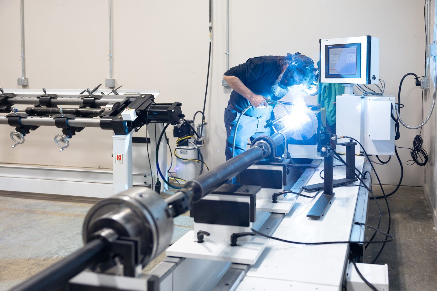 A person welds a metal driveshaft in a workshop, sparks flying. A machine and monitor are nearby.