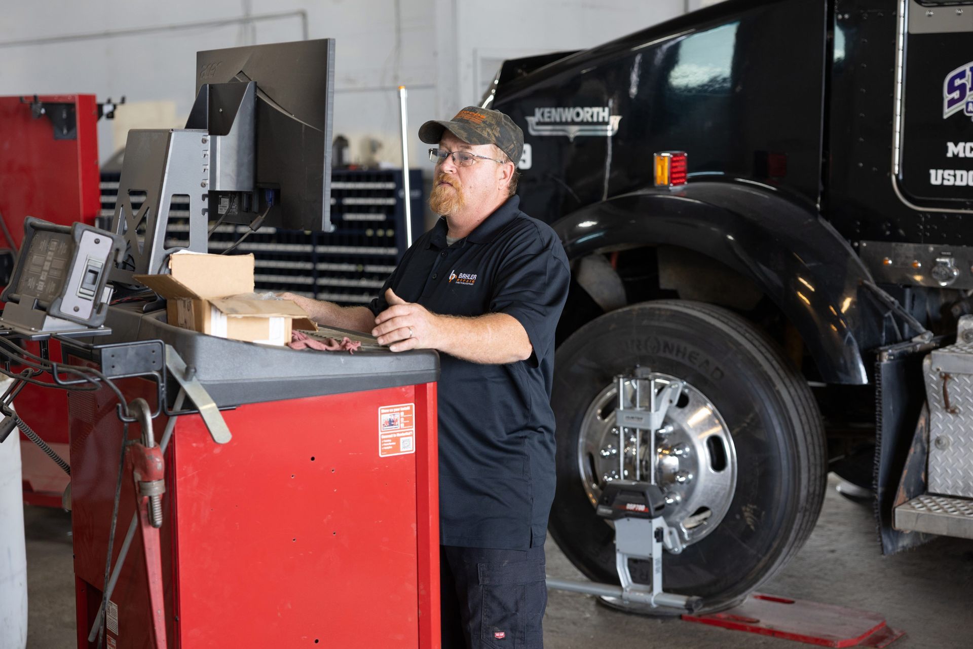Mechanic in a black shirt and hat stands next to a red machine, working on a truck tire in a garage.