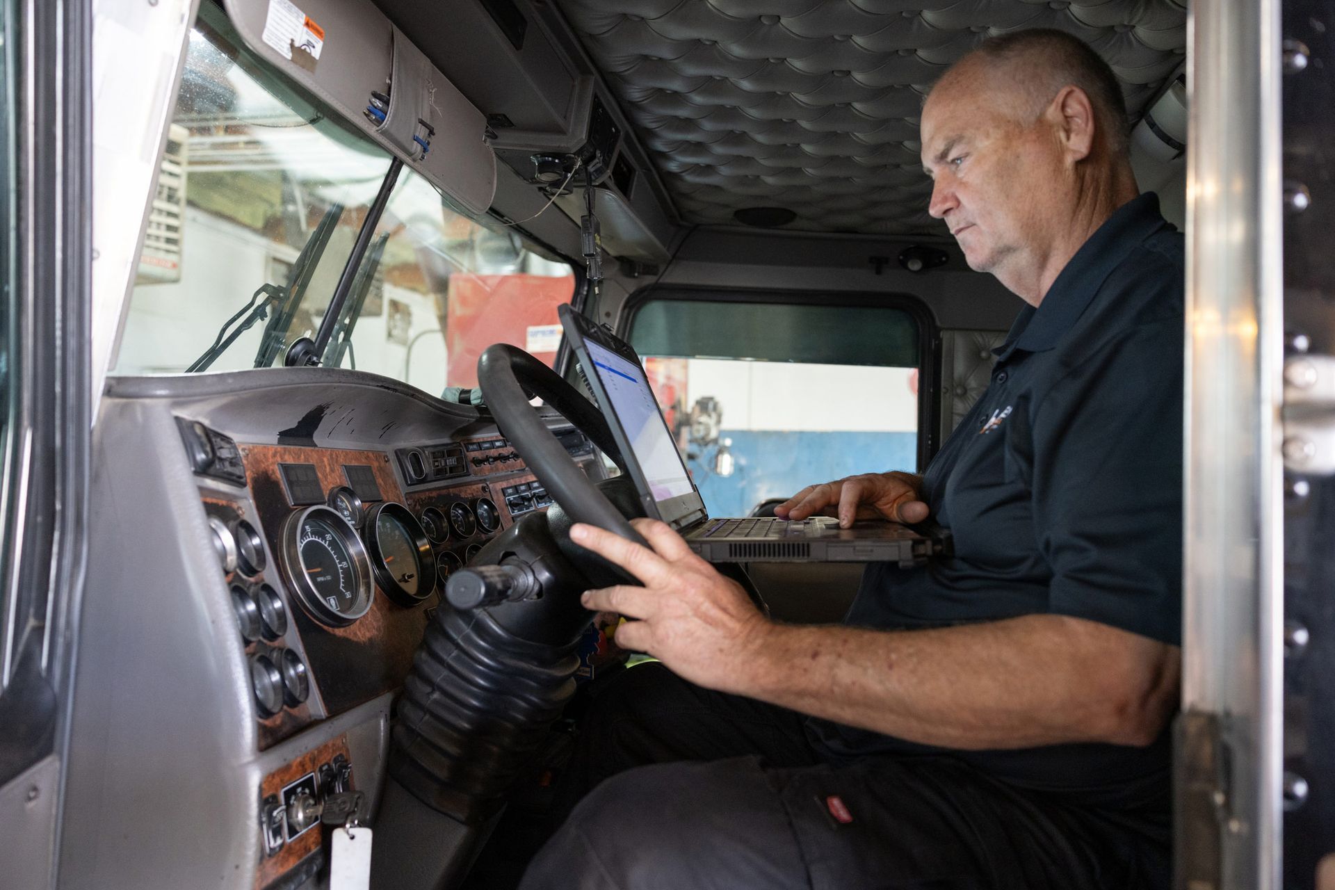 A bald man sits in the driver's seat of a semi-truck, using a laptop. He's inside the cab, working on a keyboard.