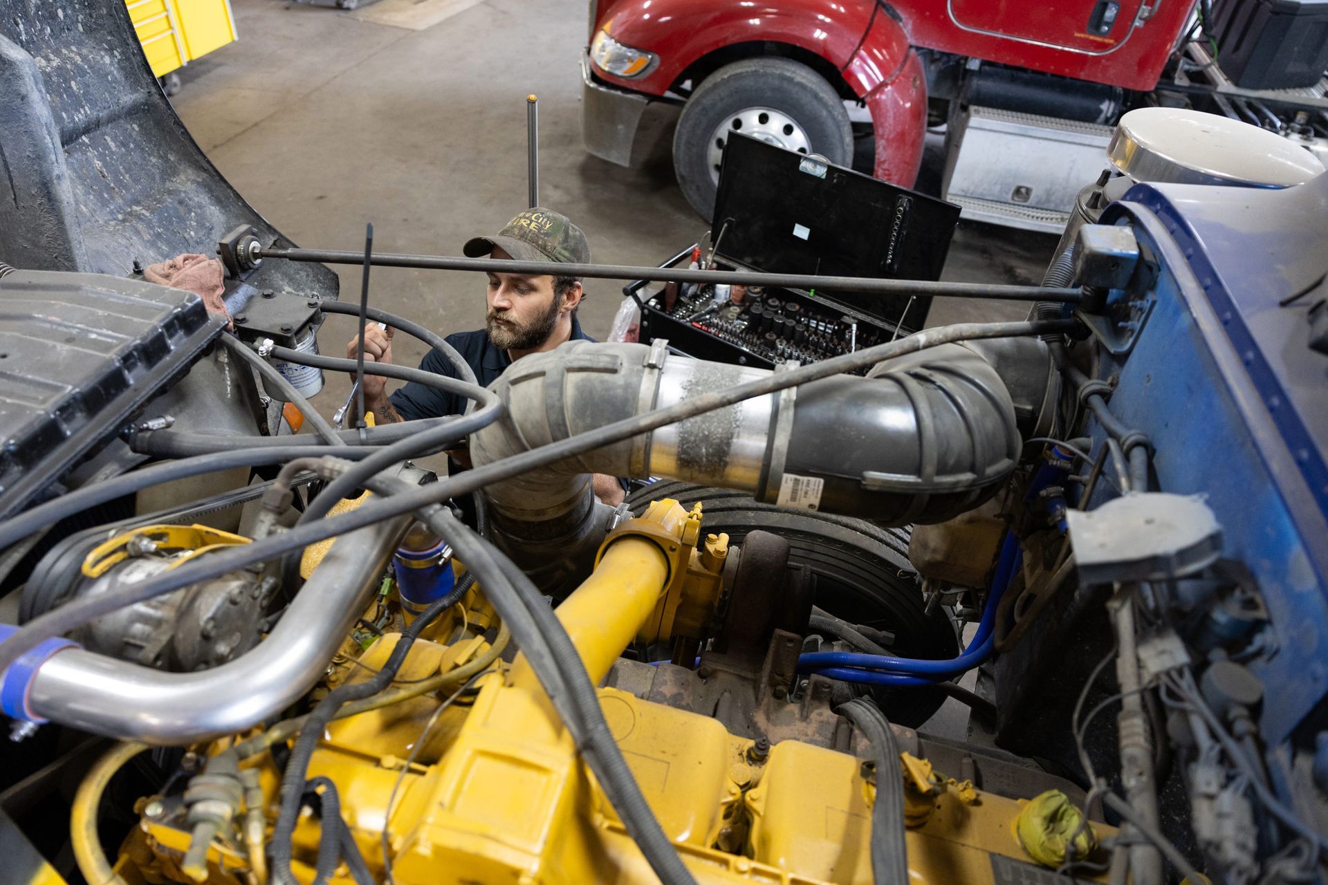 Mechanic working on a large truck engine, visible from the front. The mechanic is wearing a cap and using tools in a garage setting.