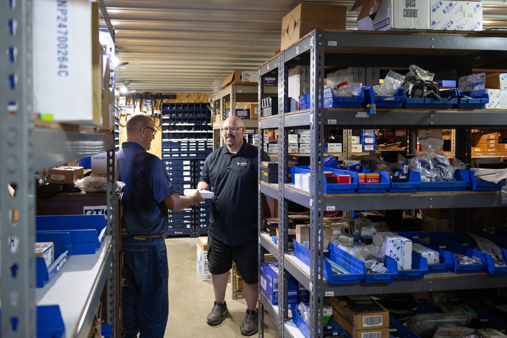 Two men in a warehouse with metal shelving filled with parts. One man is holding something while facing the other, smiling.