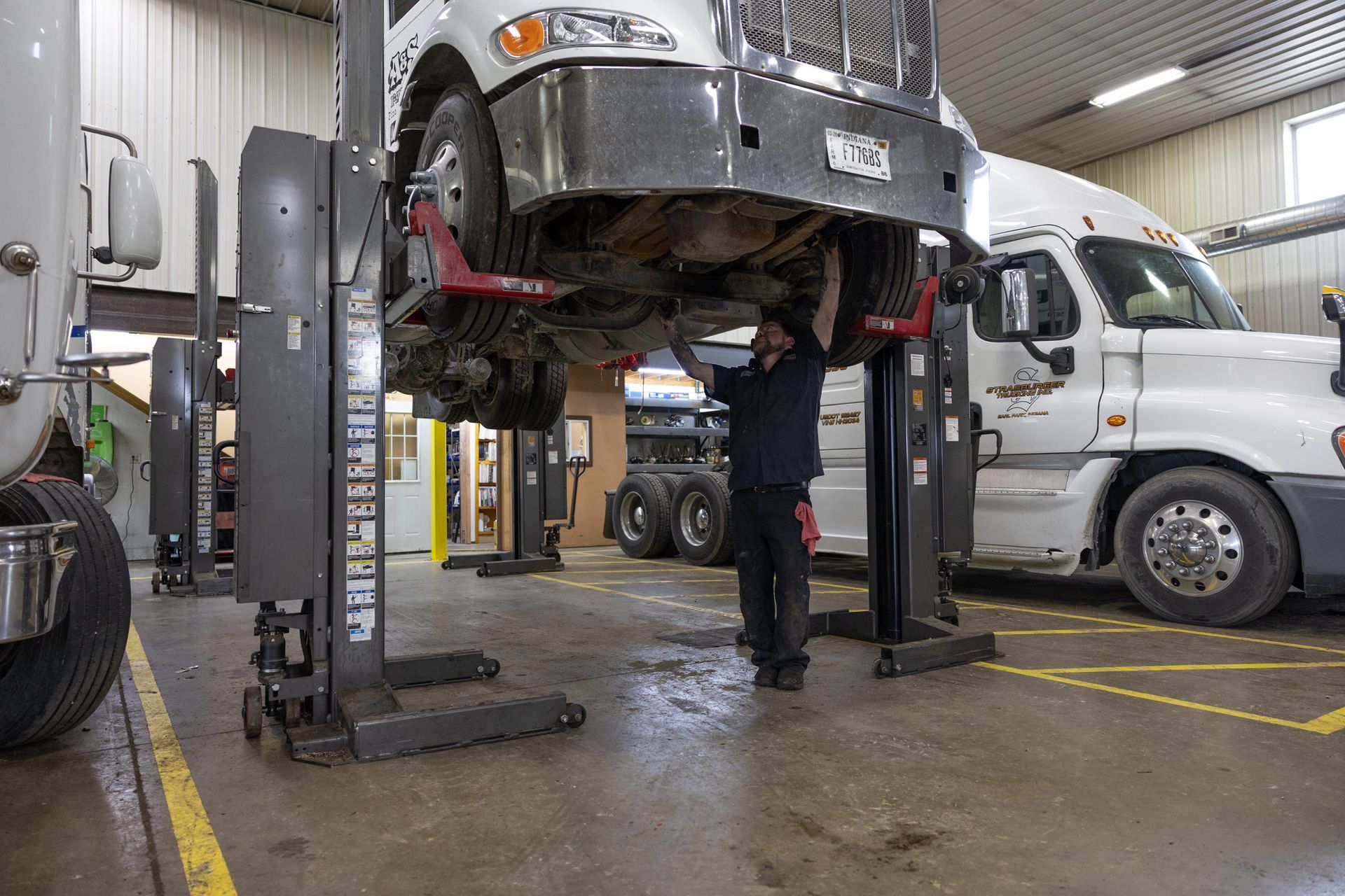 A mechanic works under a lifted semi-truck in a repair shop. Other trucks and equipment are visible.