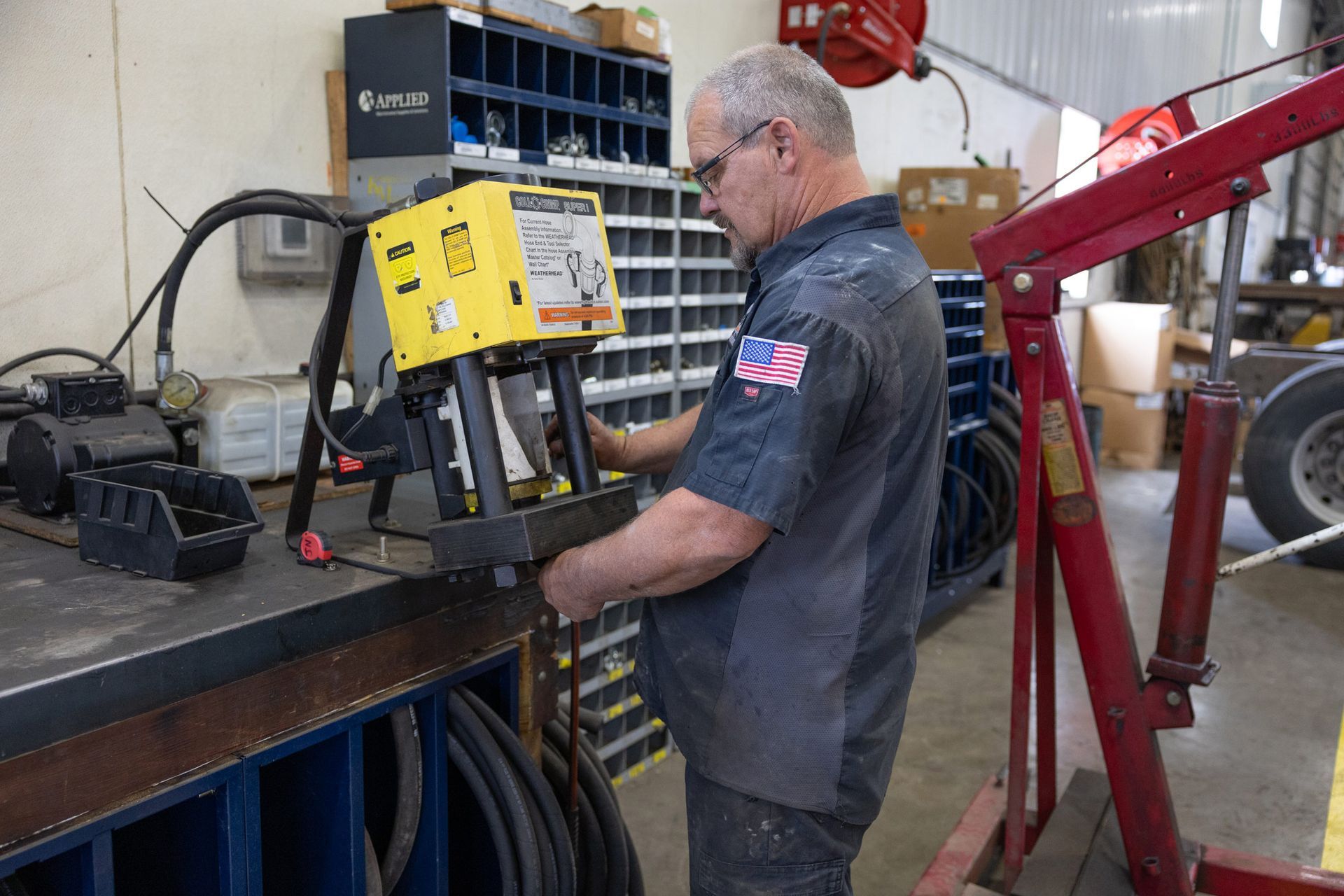 A mechanic works on metal with a yellow tool in a workshop. He wears glasses and a shirt with a US flag patch.