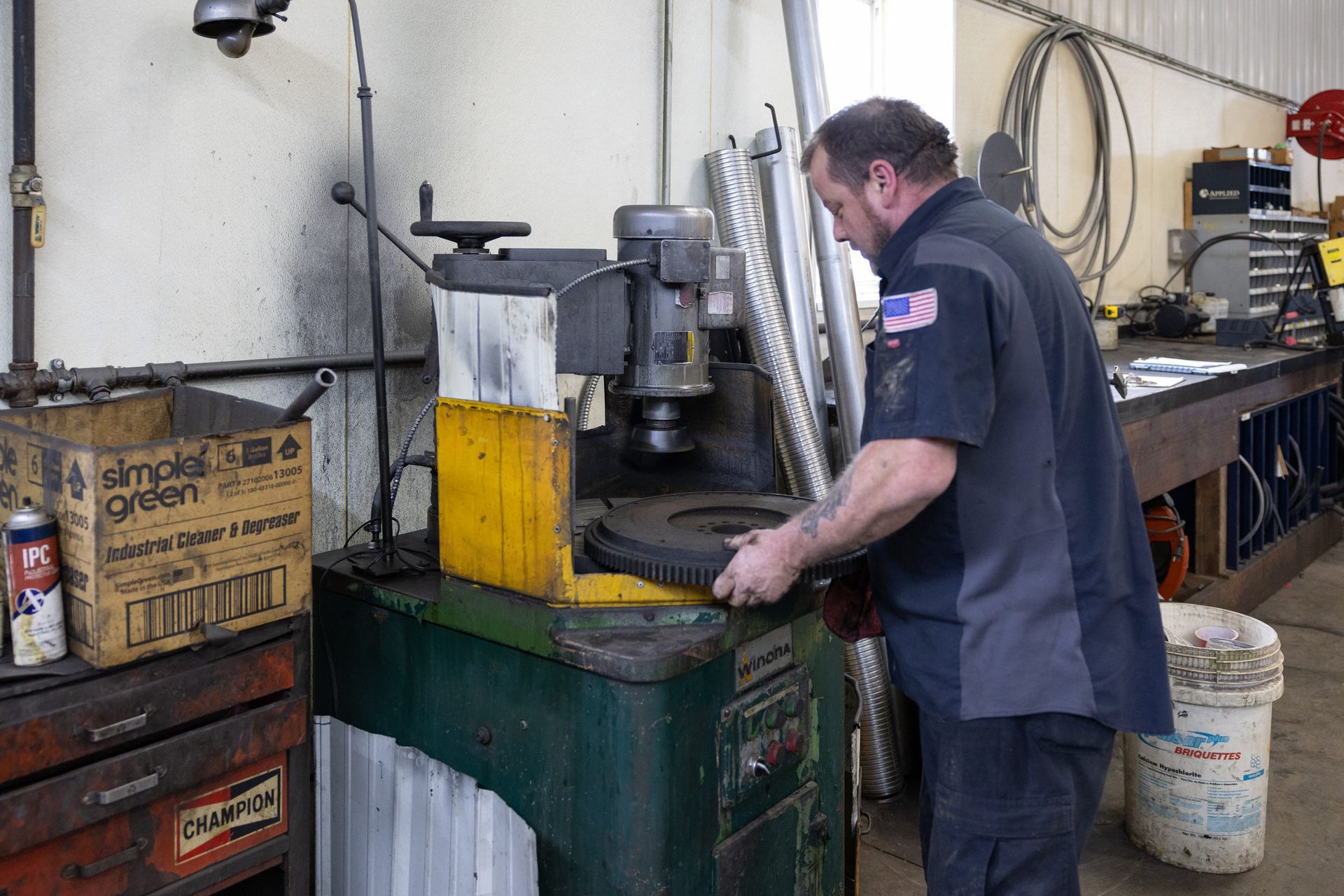 A mechanic working on machinery in a shop. He is holding a large, dark, circular part over a press.