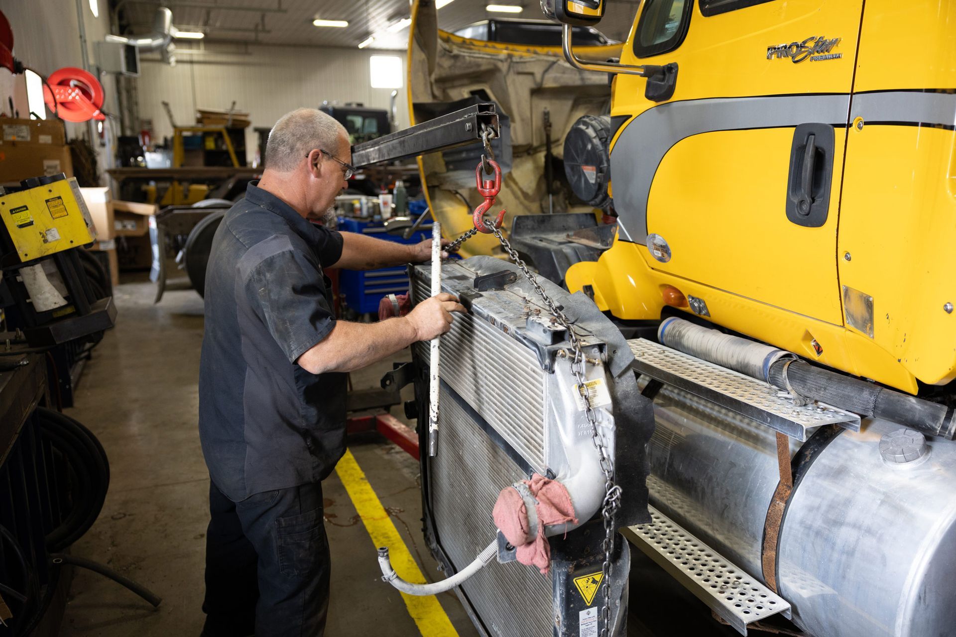 Mechanic removing a radiator from a yellow truck in a repair shop. The mechanic is wearing a grey shirt and using a crane.
