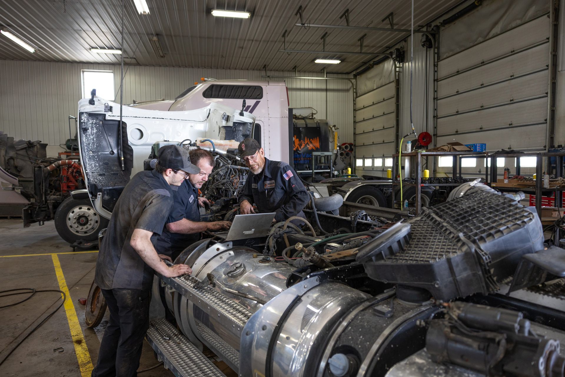 Three mechanics working on a truck engine in a garage. They examine the engine, one using a laptop.