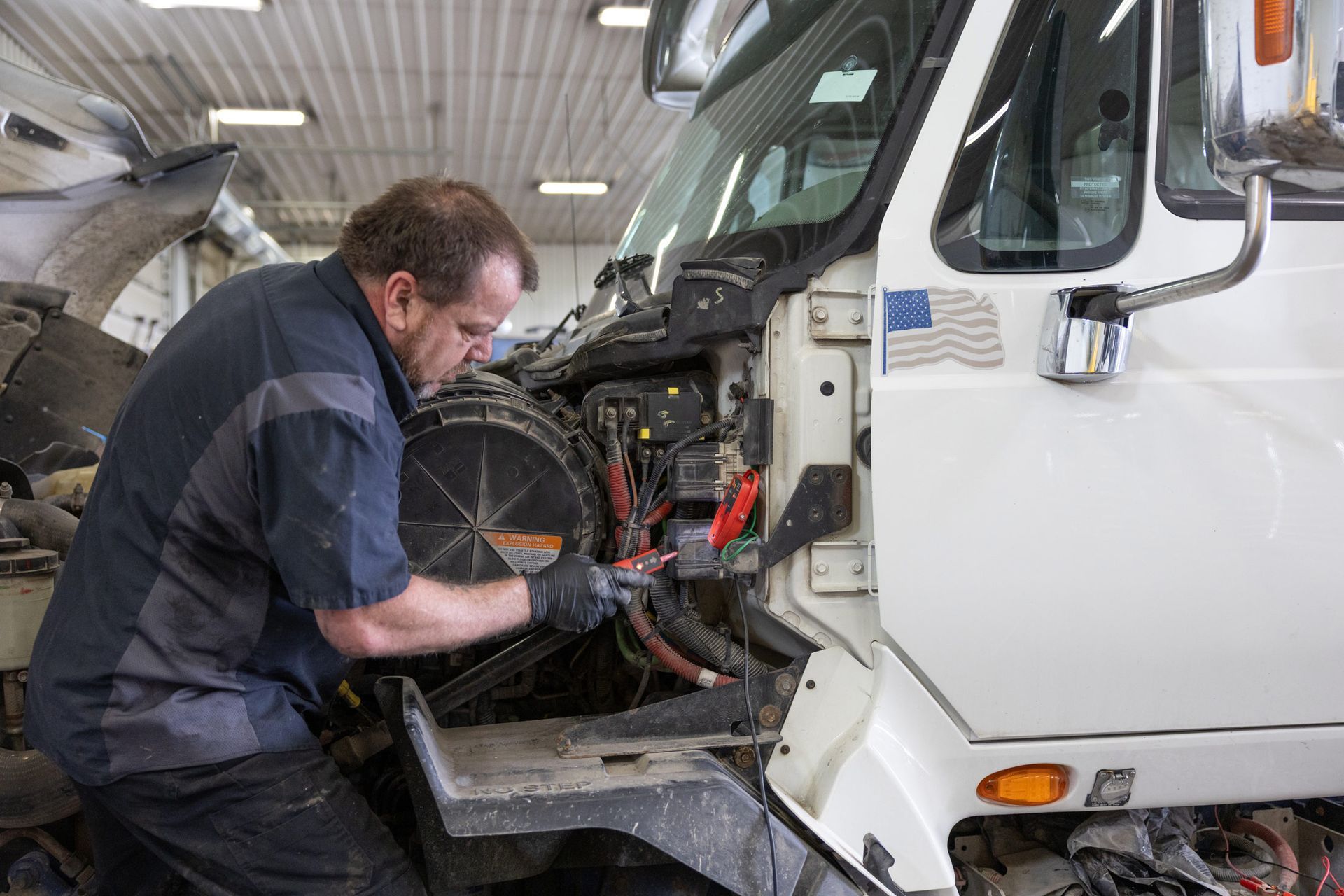 Mechanic in a garage repairs a white truck, focusing on its engine. He wears gloves, and the setting is bright.