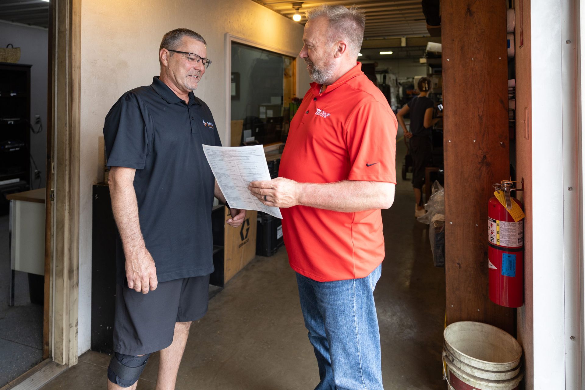 Two men, one in a red shirt and the other in black, talking near a doorway, holding papers. They are in a garage-like space.