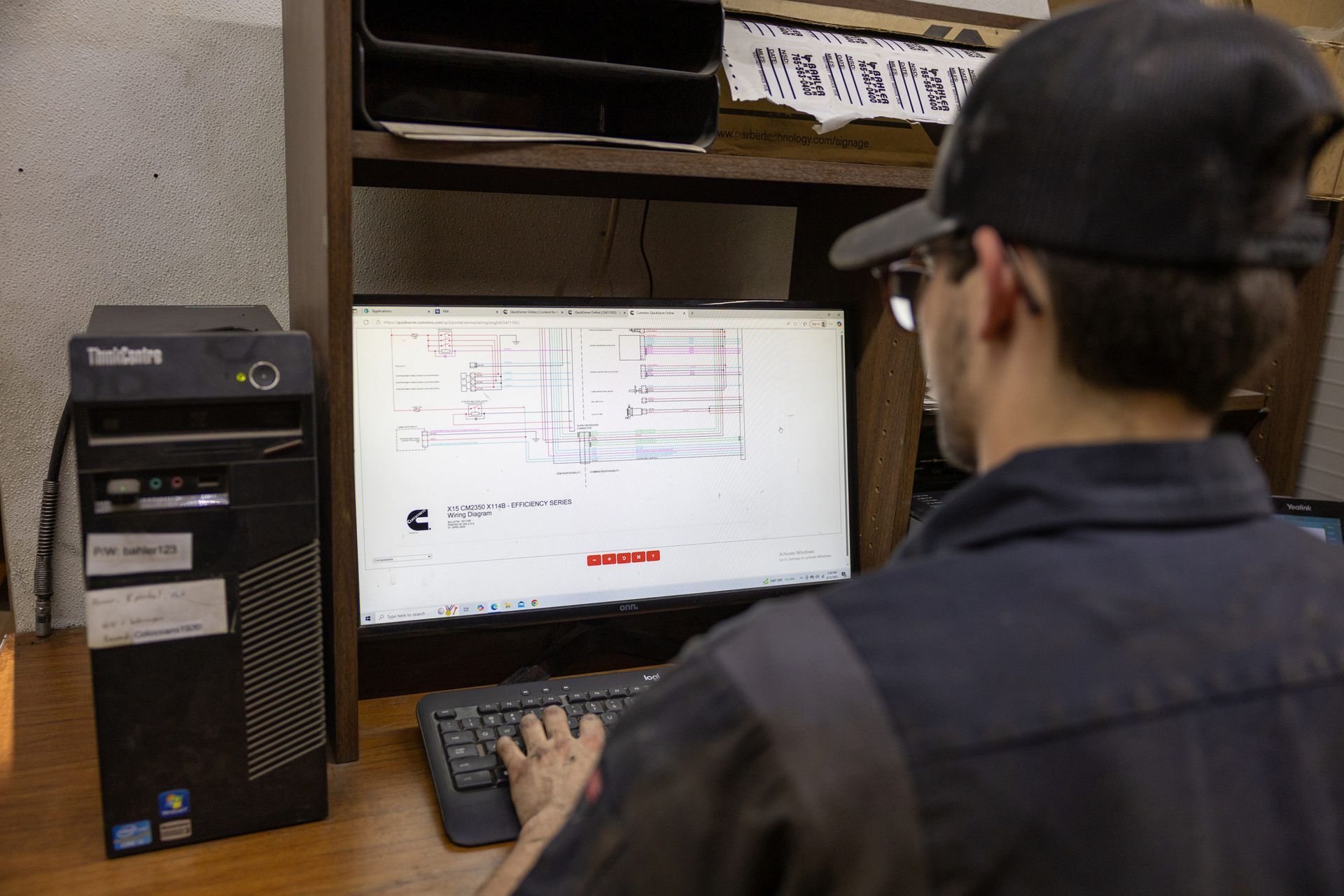 Person in work clothes using a computer, likely reviewing blueprints. A desktop computer is beside the monitor in a workshop setting.