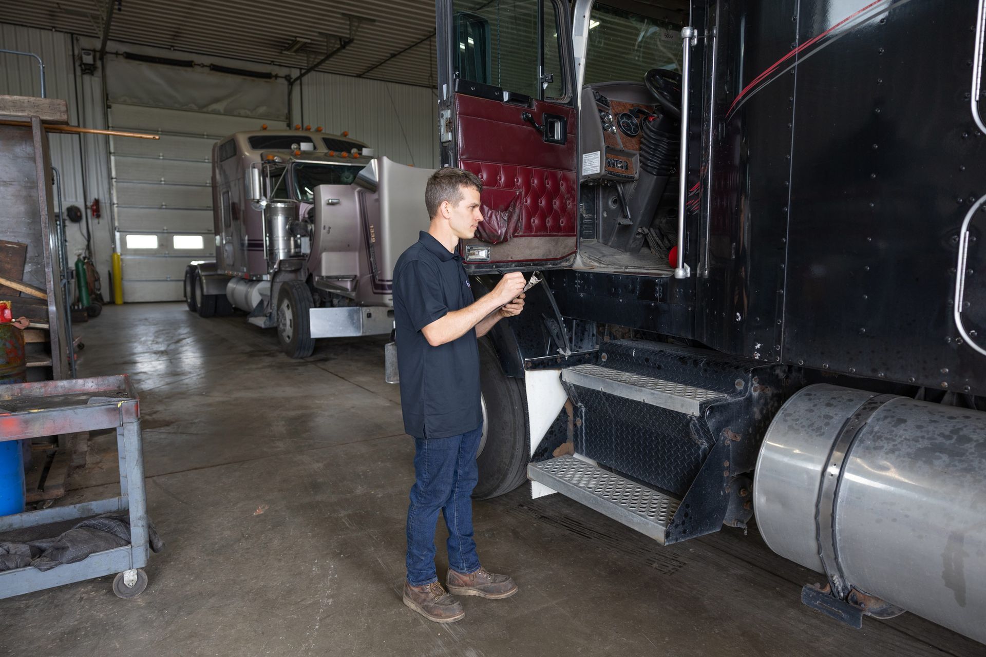 Mechanic inspects black semi-truck in a repair shop, another truck in the background. He wears a dark uniform and holds a tool.