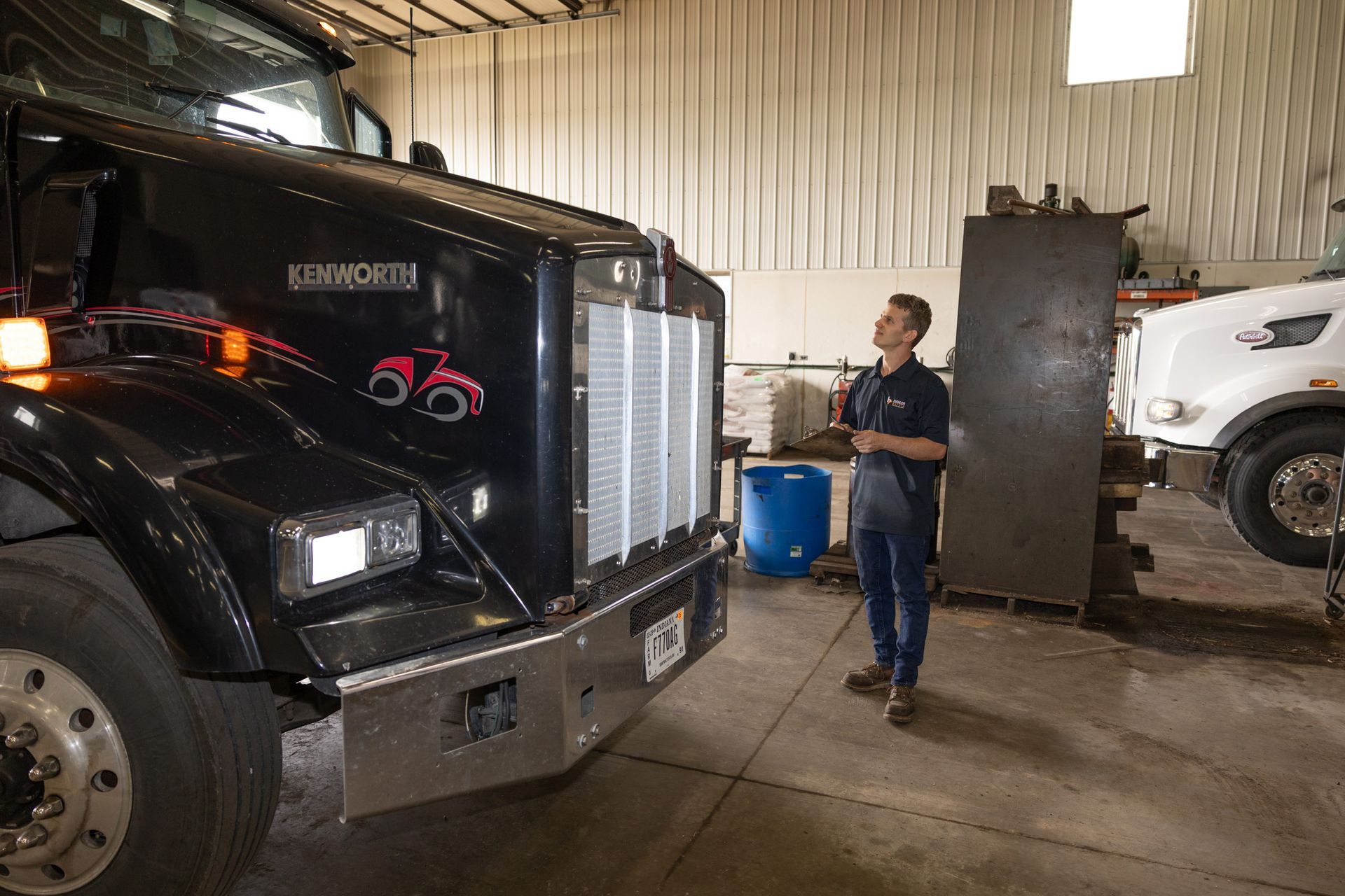 A mechanic inspects a black Kenworth truck in a garage. A second white truck is visible in the background.
