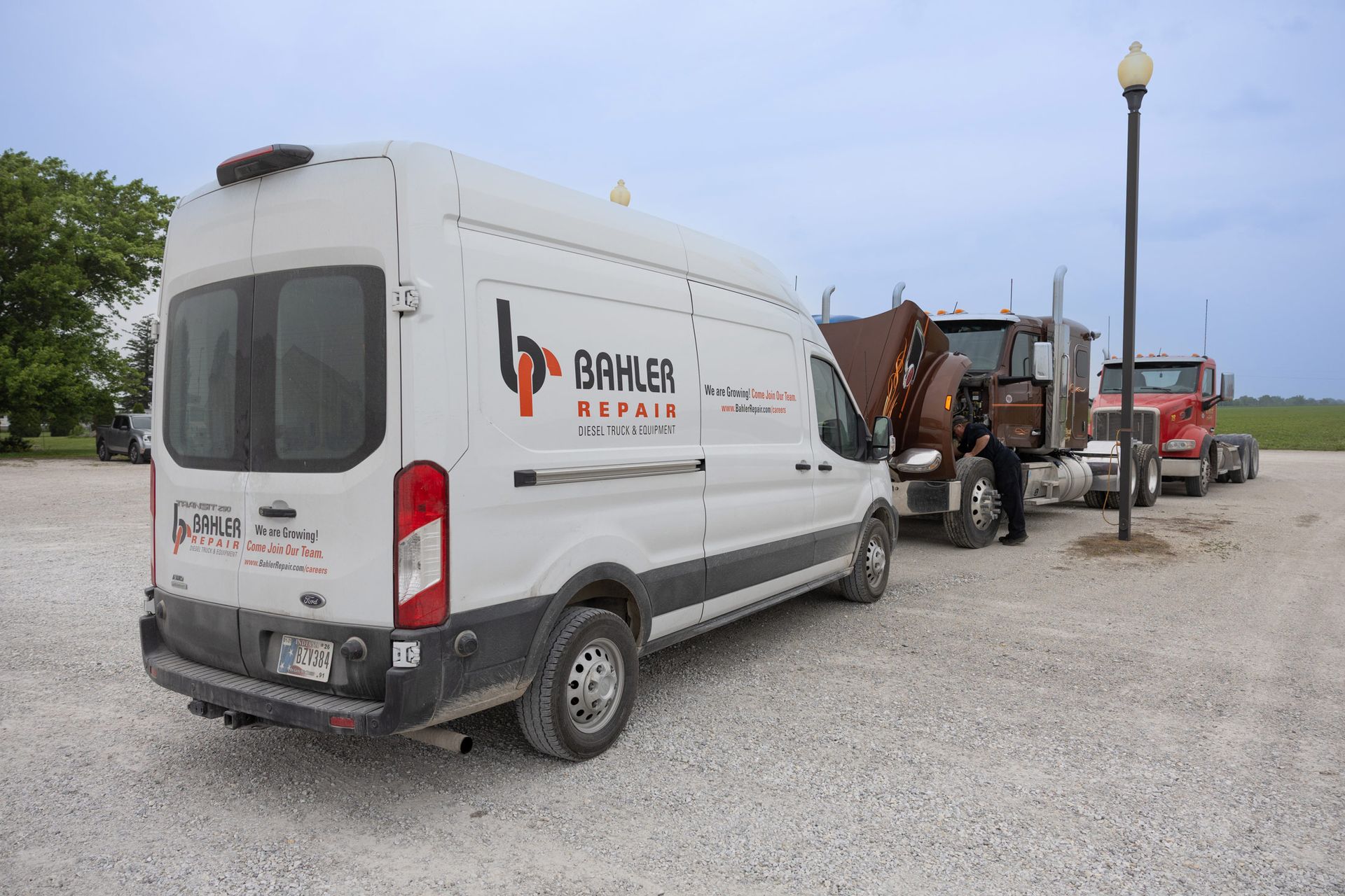 White service van parked next to two semi-trucks on gravel. Person in dark clothing stands near the trucks in an outdoor setting.