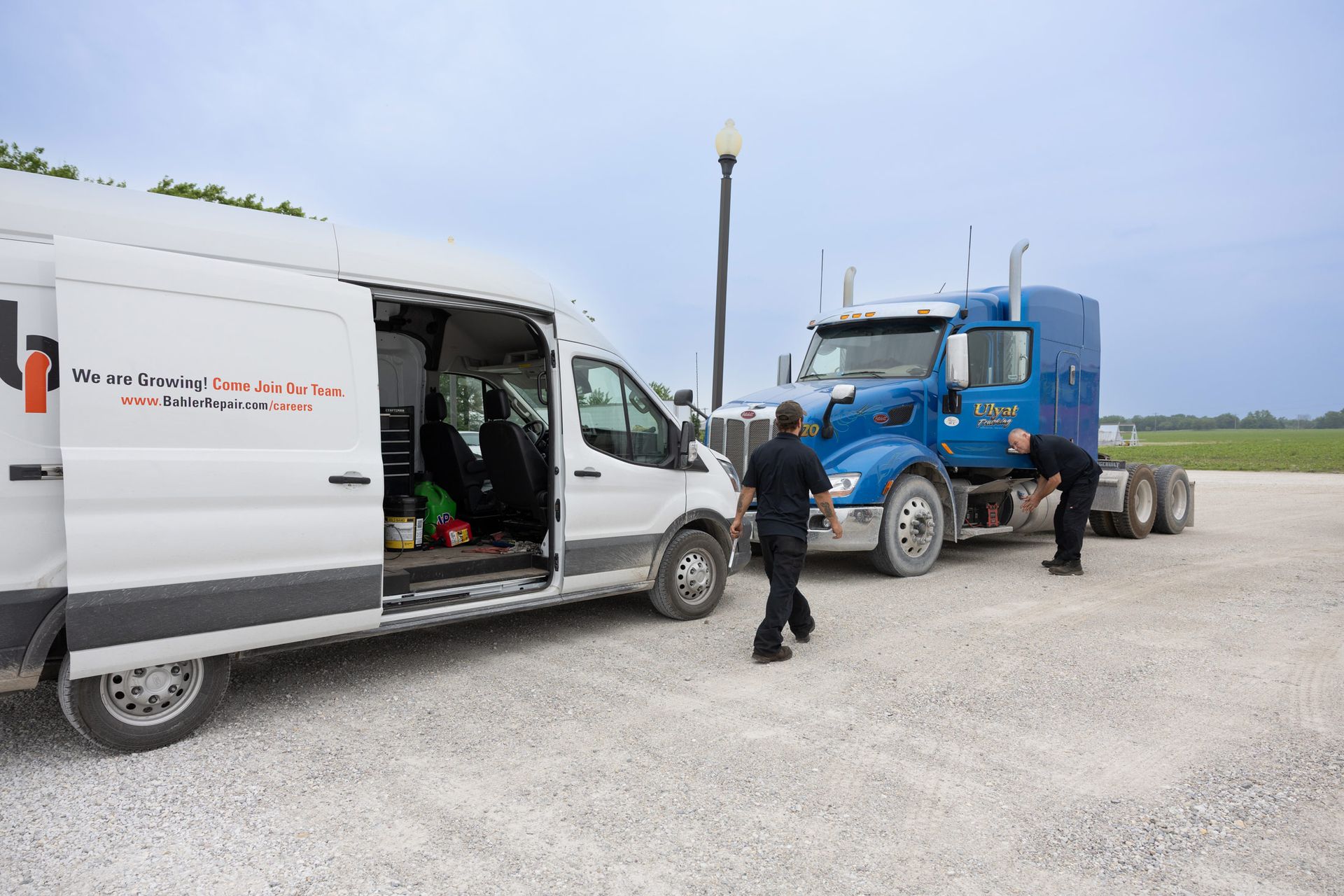 A white service van is parked beside a blue semi-truck, with two people inspecting the vehicles on a gravel road.