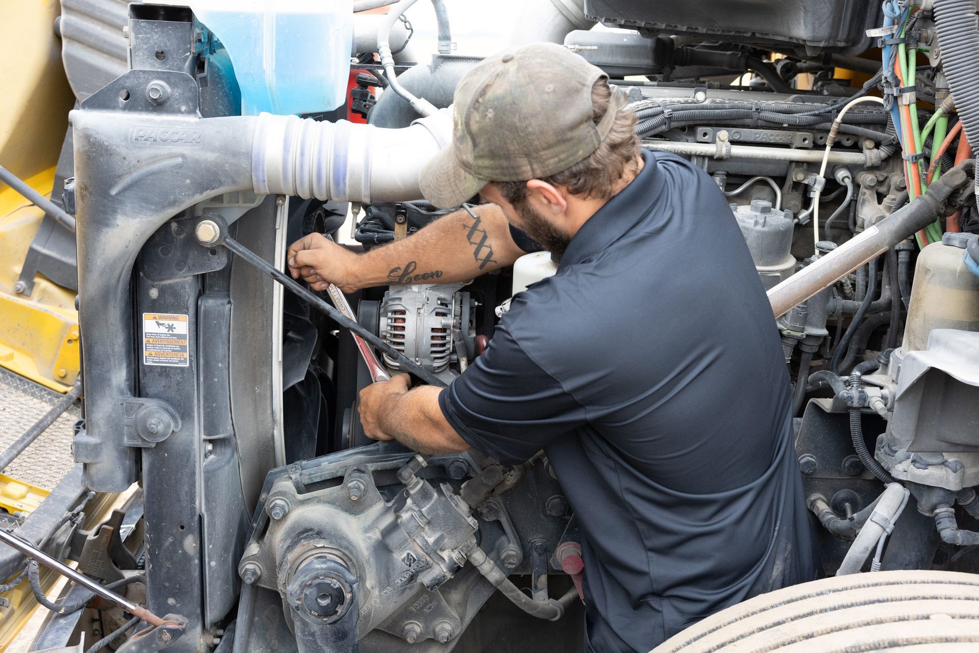 Mechanic in a black shirt and cap working on a truck engine, holding a wrench.