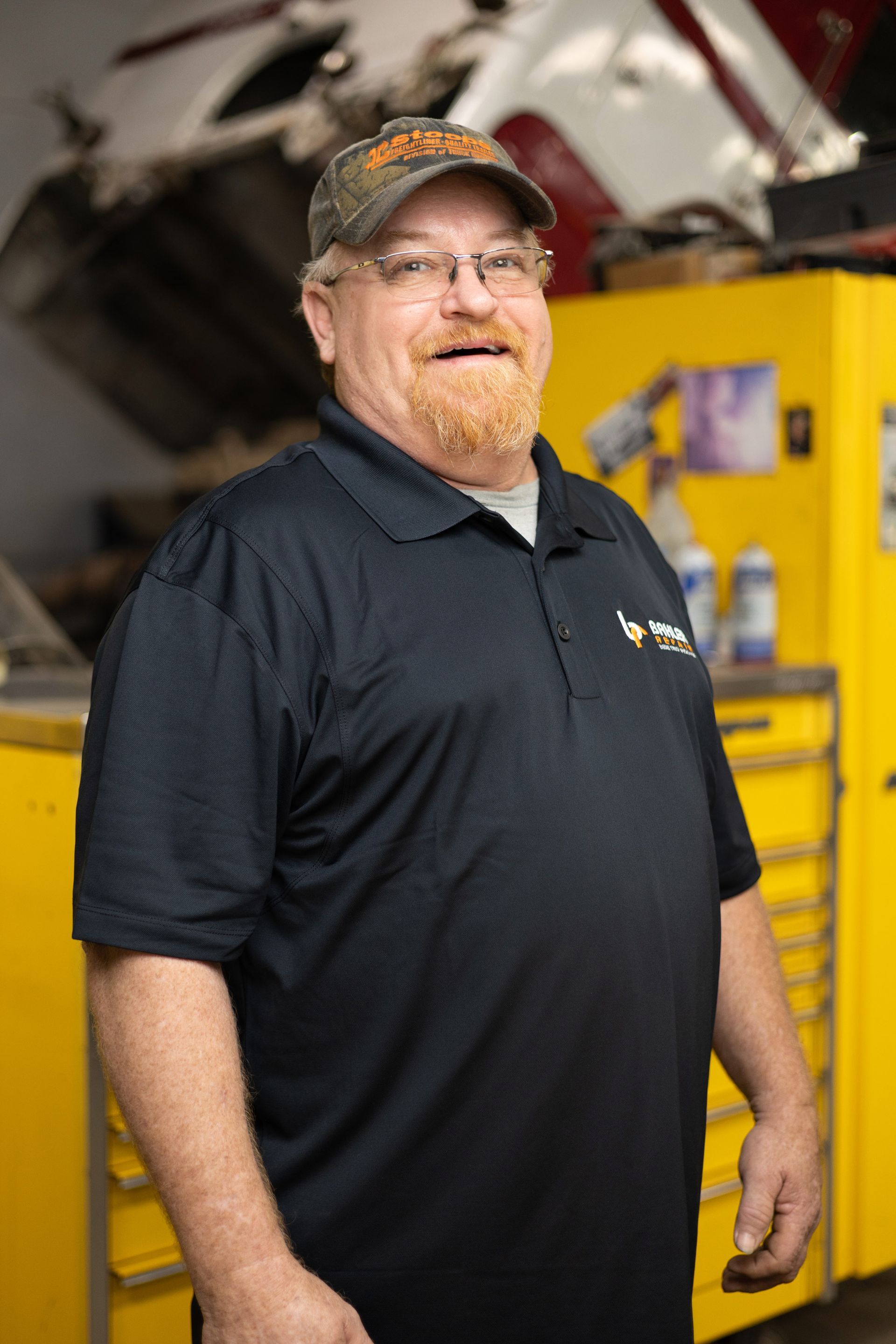 Man with a beard wearing a baseball cap and polo shirt, smiling in an auto repair shop.