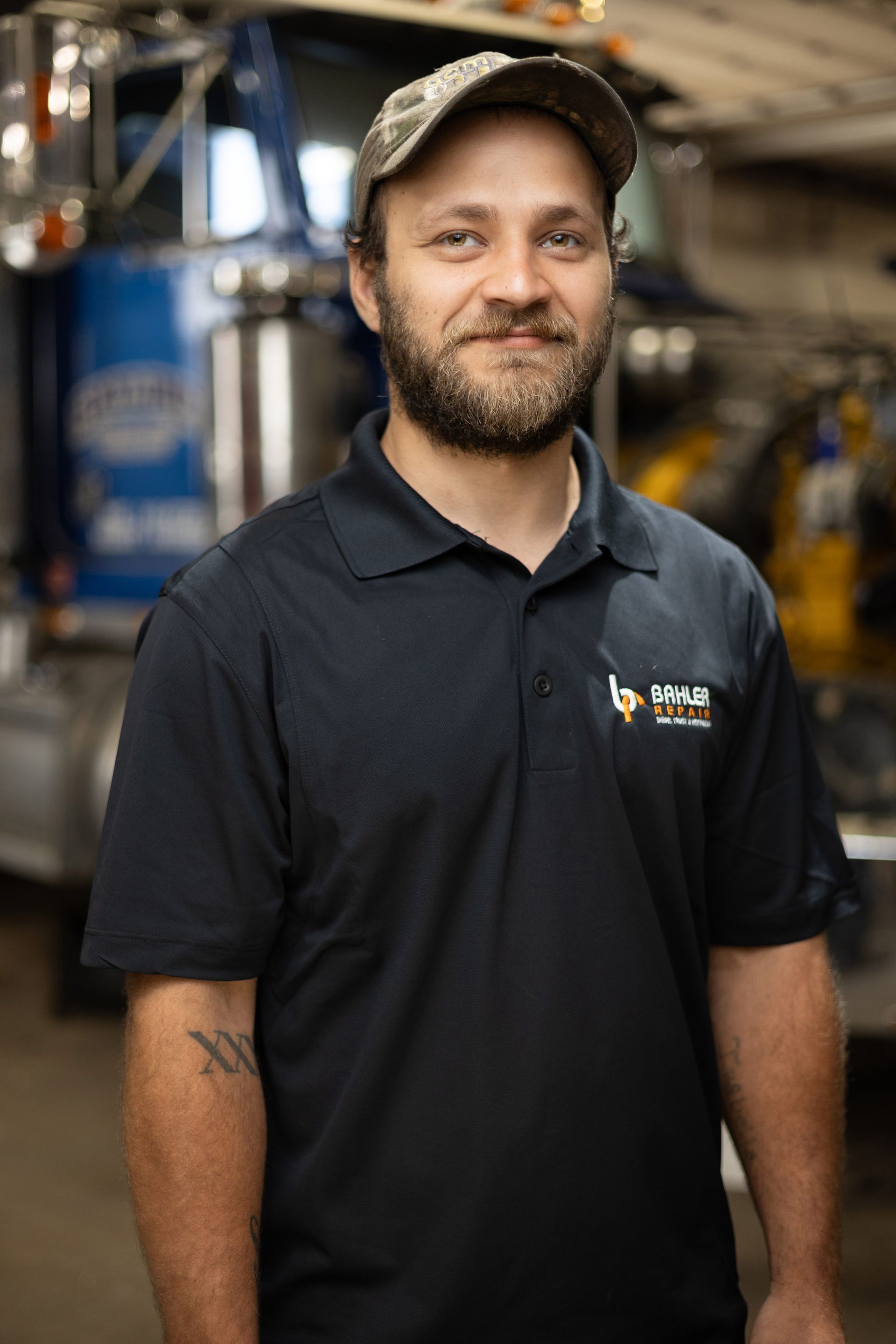 Man with beard and baseball cap in a dark polo shirt smiles in front of a truck, presumably in a mechanic shop.