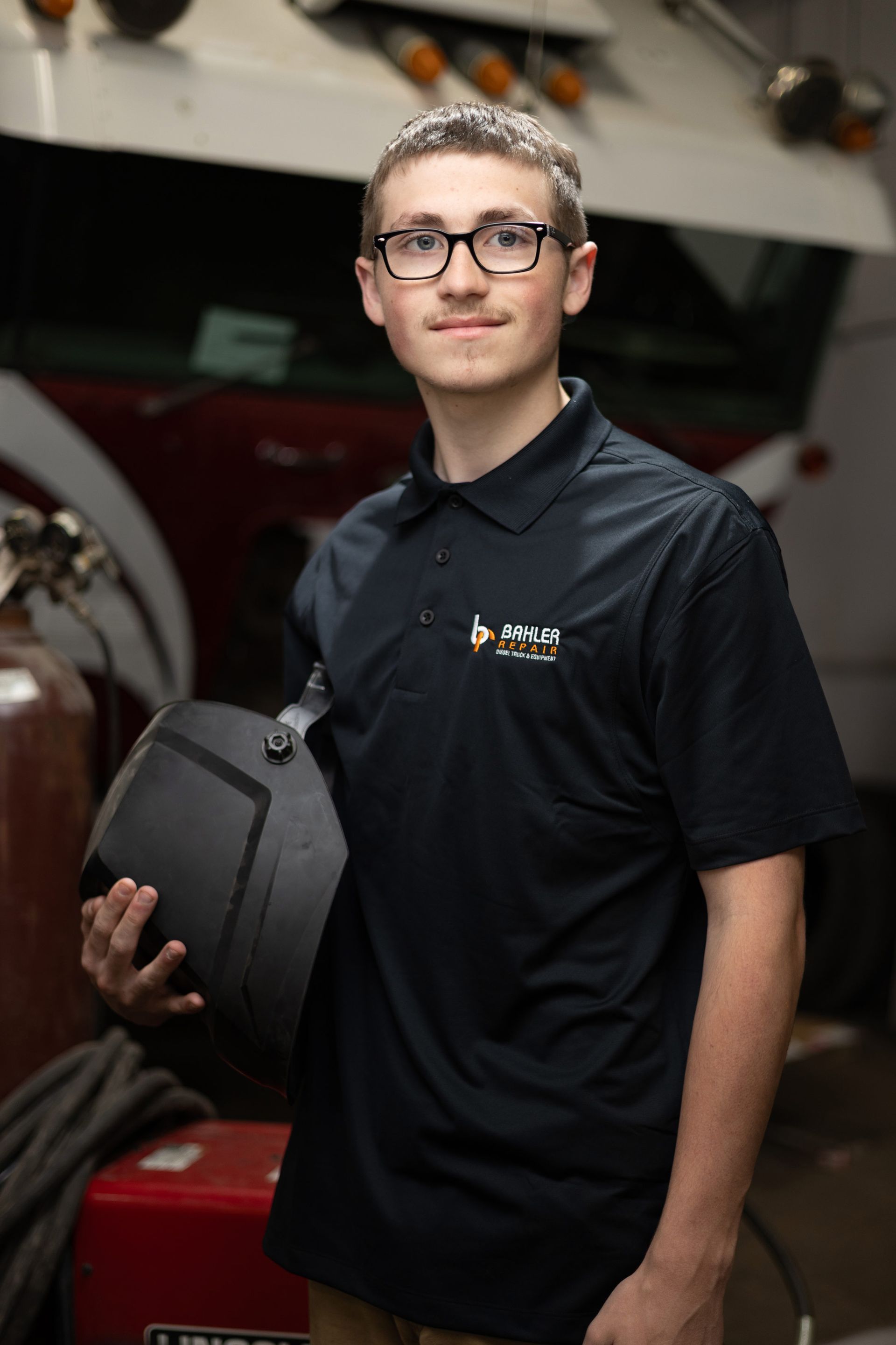 A young man in a black polo shirt holds a welding helmet, posing in a workshop setting. He wears glasses and smiles.