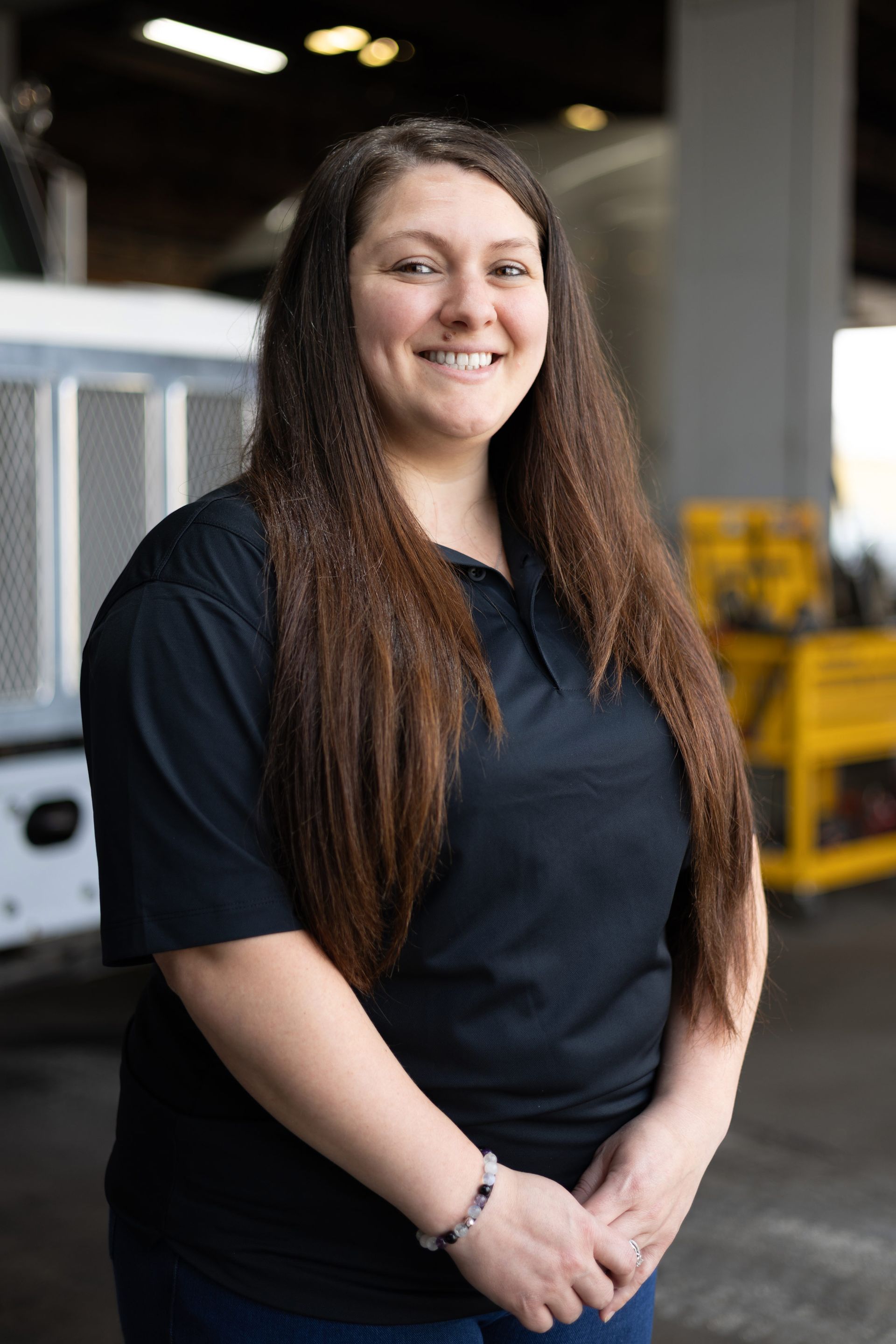 Woman with long brown hair, wearing a black shirt and jeans, smiling in a garage setting.