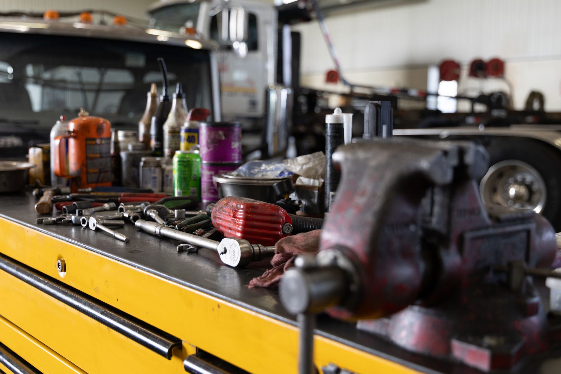 A cluttered workbench in a garage with tools, bottles, and a red vise. A semi-truck is visible in the background.