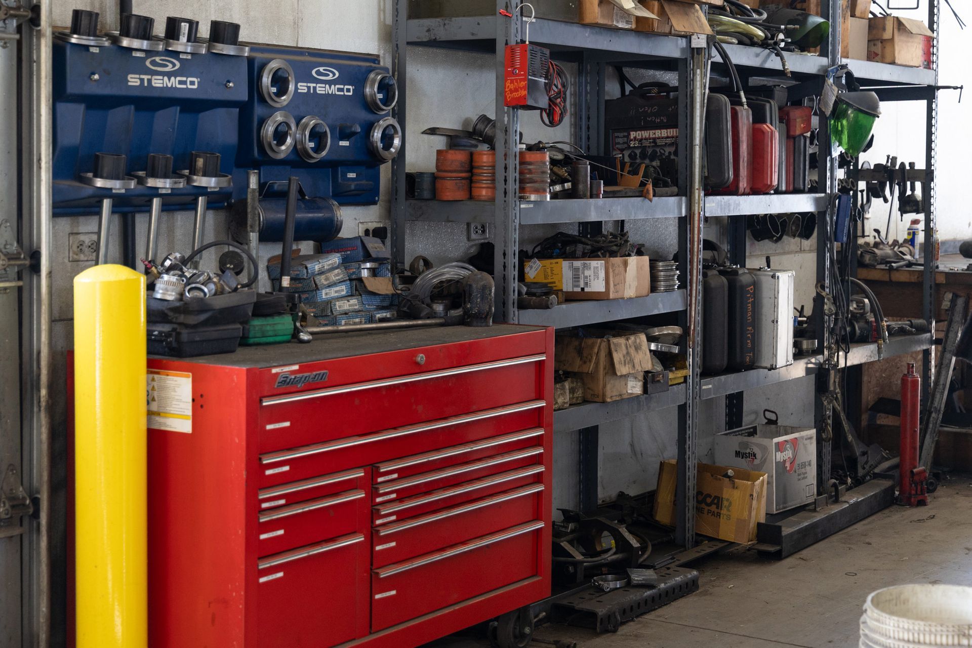 Red tool chest and yellow post in a cluttered mechanic's shop. Shelves hold tools and parts.