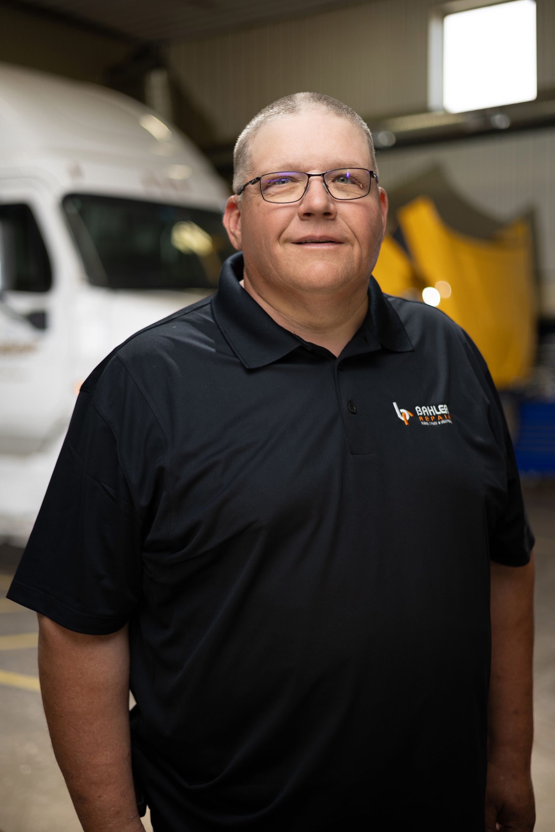 Man wearing glasses and a black polo shirt stands in a vehicle maintenance shop, a white truck is in the background.