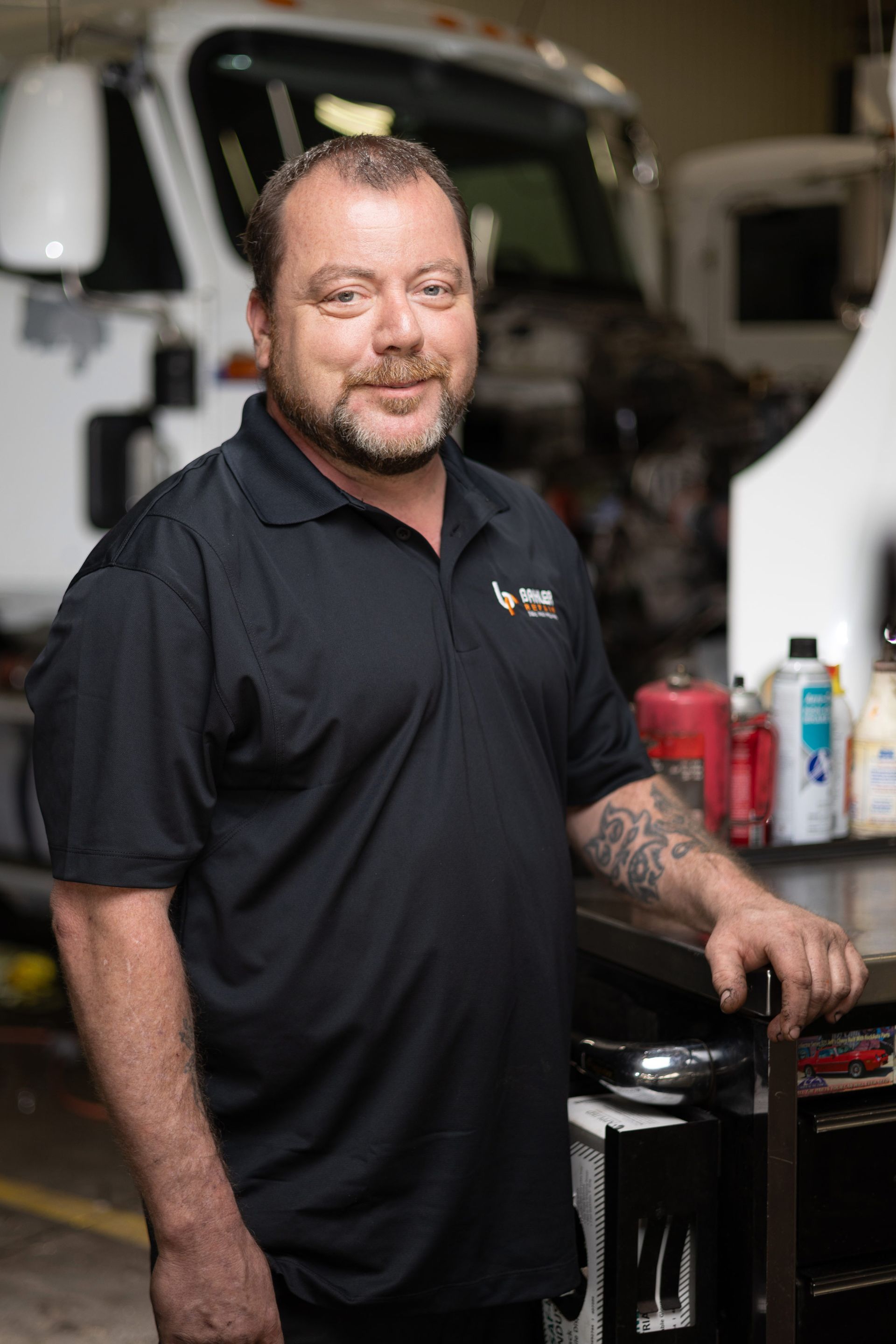 Man in black shirt with arm tattoos, smiling in a mechanic shop, standing by a tool cart. A truck is visible in the background.