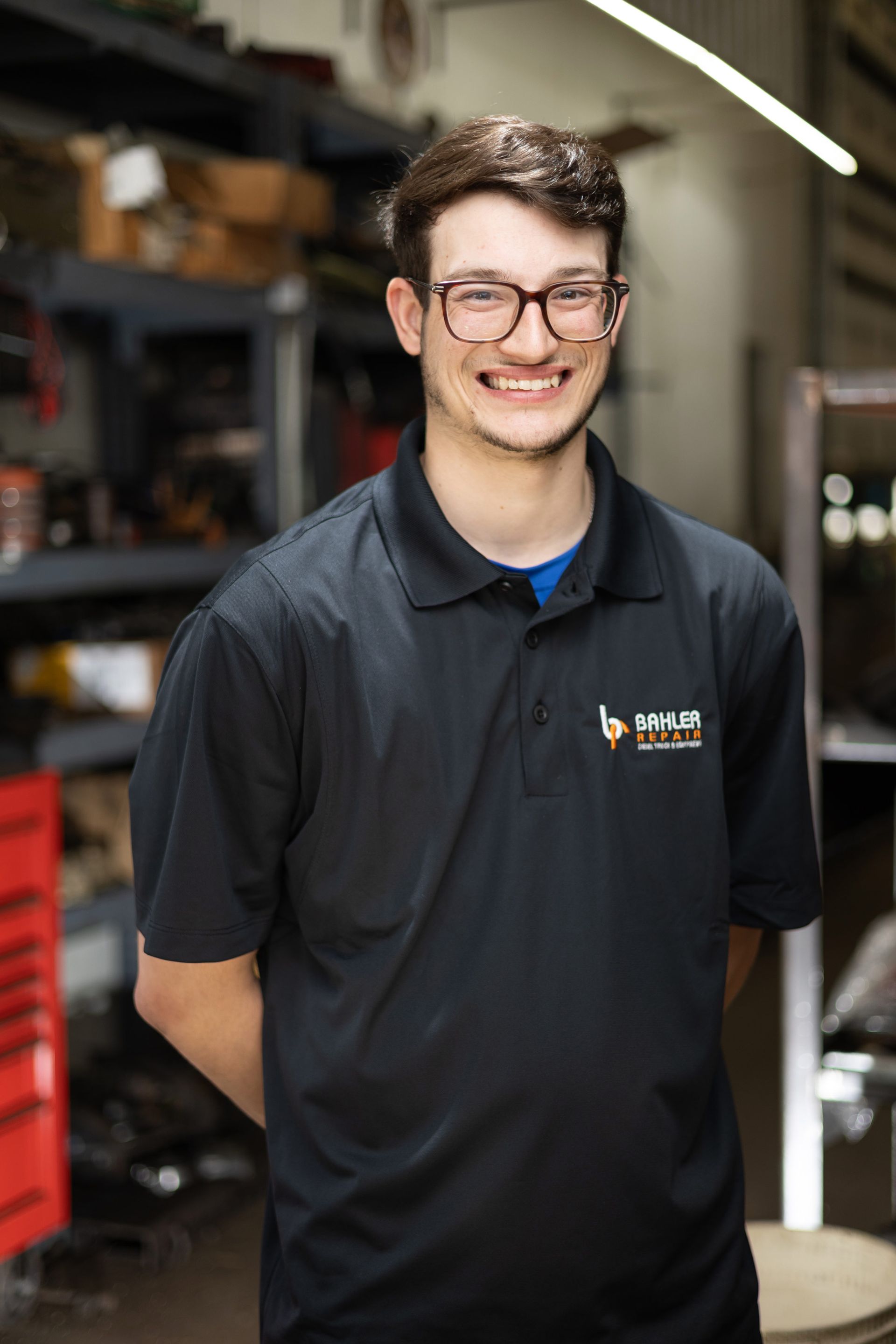 Young man with glasses and a black polo shirt smiles in a workshop setting, hands behind his back.