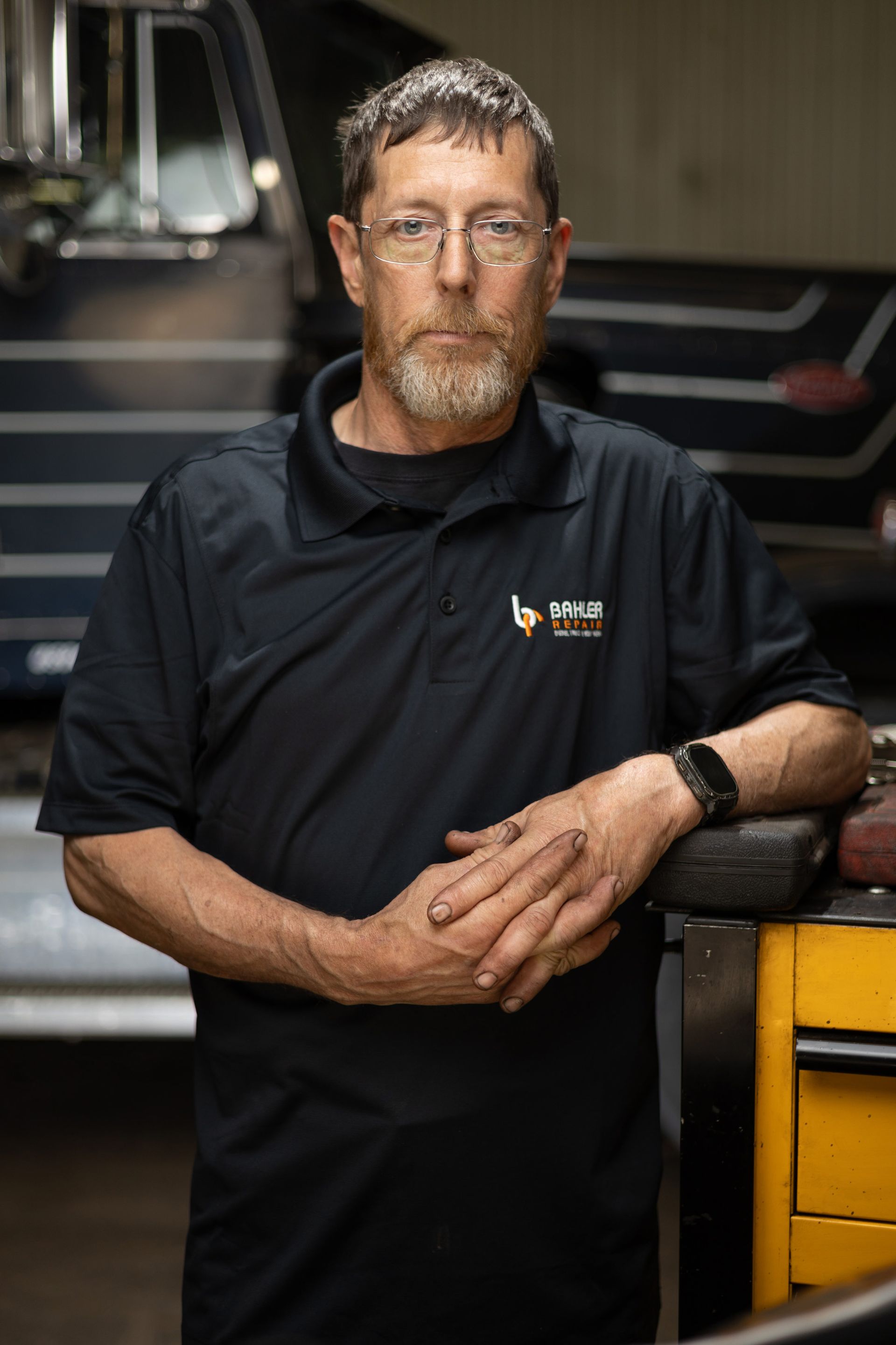 Mechanic with a beard, wearing a black polo shirt, leans on a yellow tool cabinet in a garage with a truck in the background.