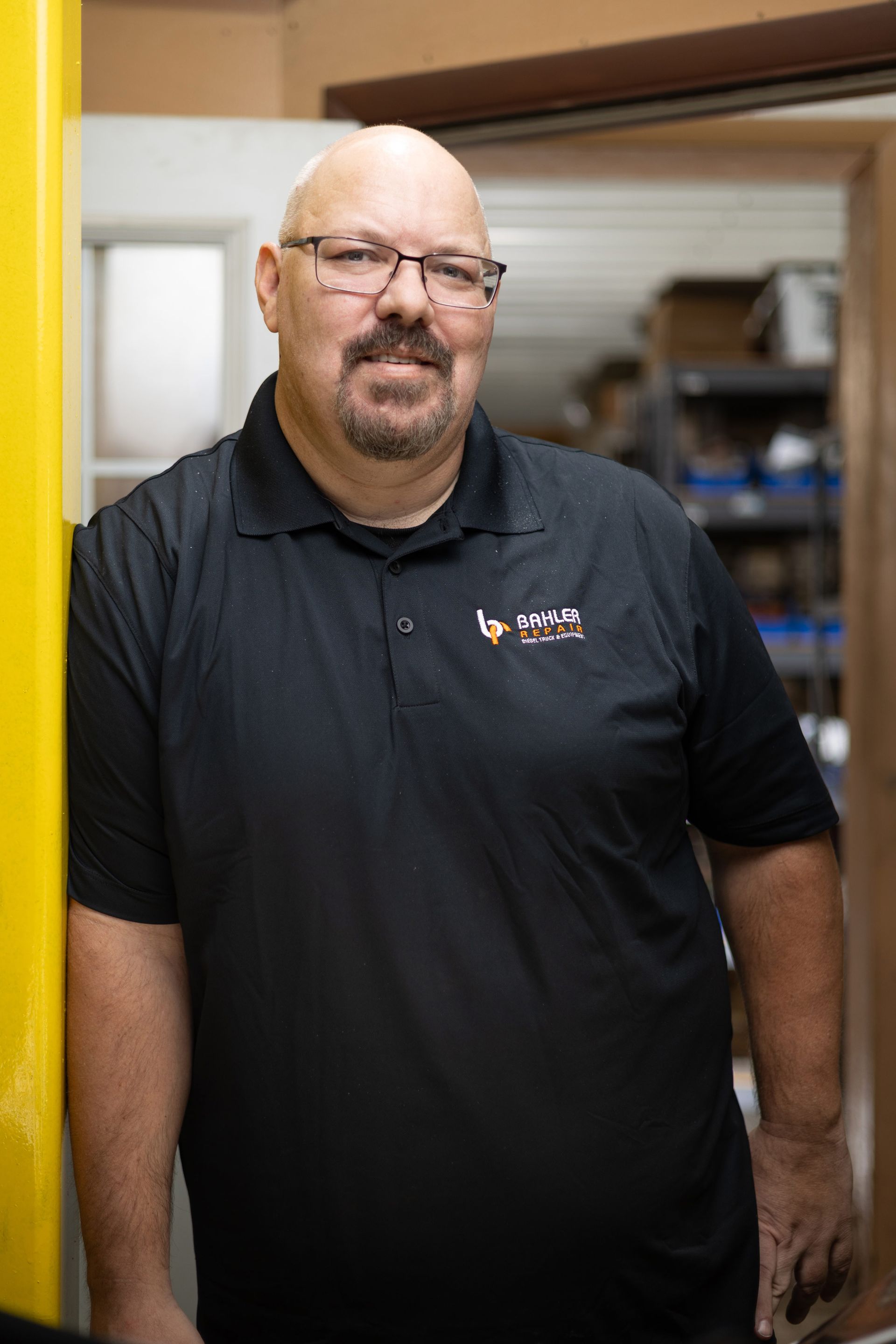 A middle-aged man with glasses and a goatee leans against a yellow pillar in an industrial setting. He is wearing a black polo shirt with a logo.