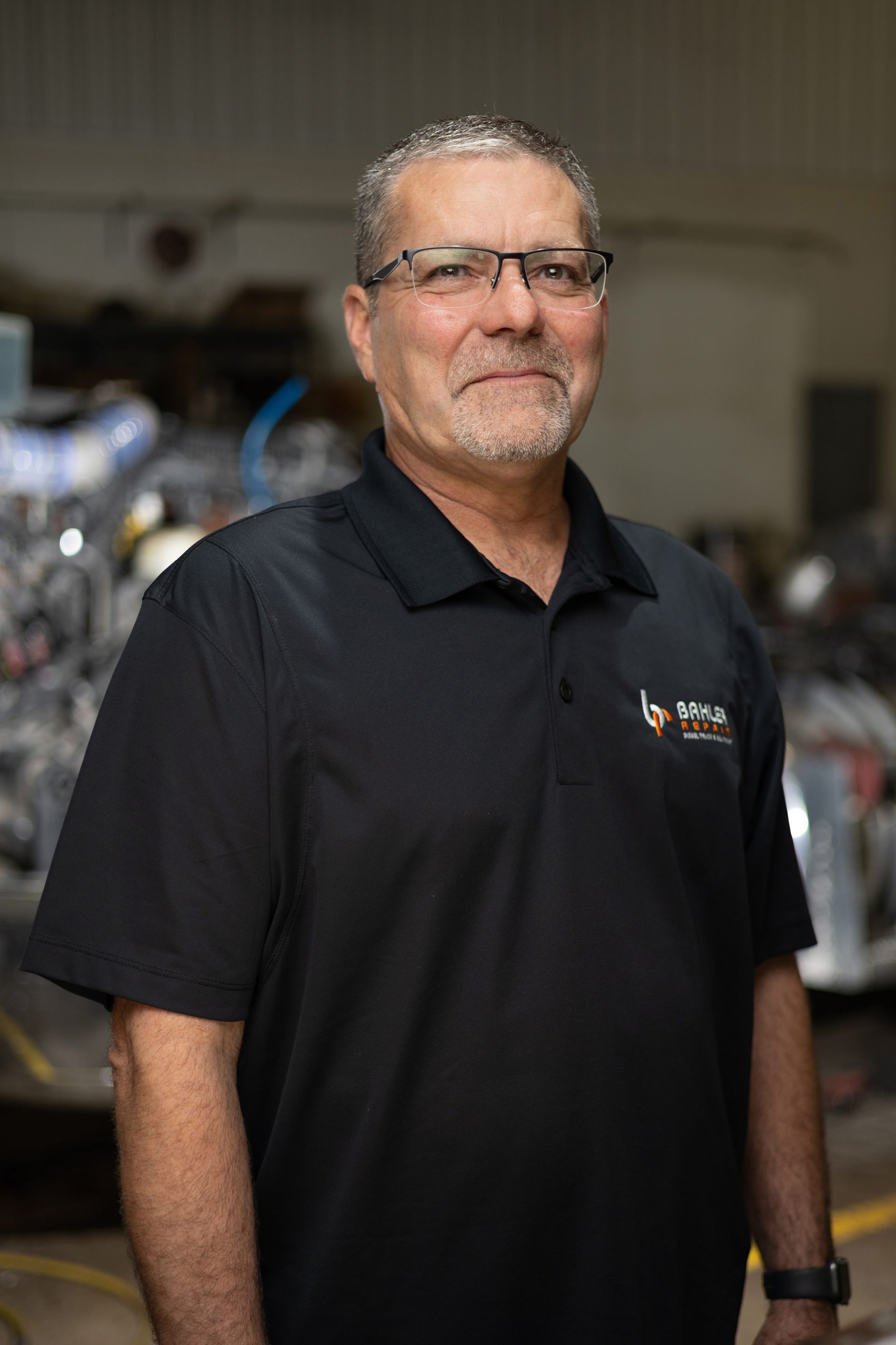 Man with gray hair and glasses in black polo shirt, smiling in a workshop setting.