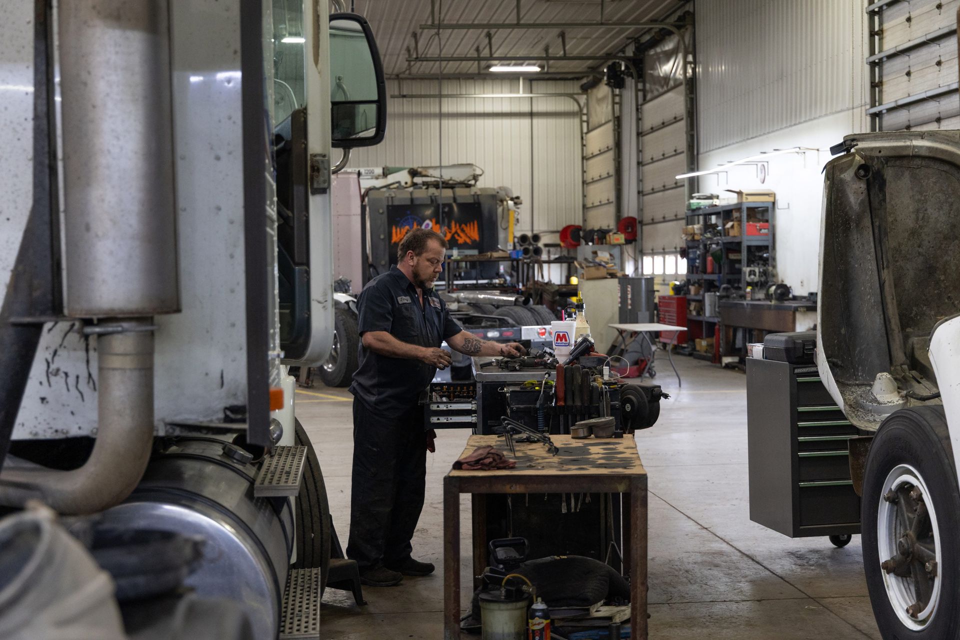 Mechanic working on machinery inside a truck repair shop. The person is standing and looking down at an object on a table.