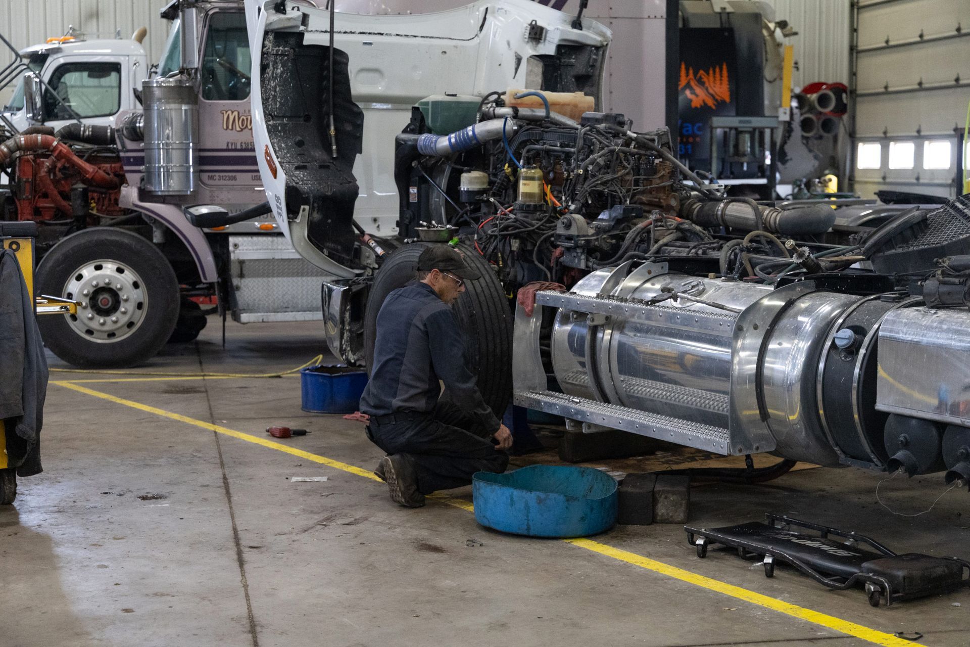 Mechanic working on a semi-truck in a repair shop, kneeling with tools and blue oil pans.