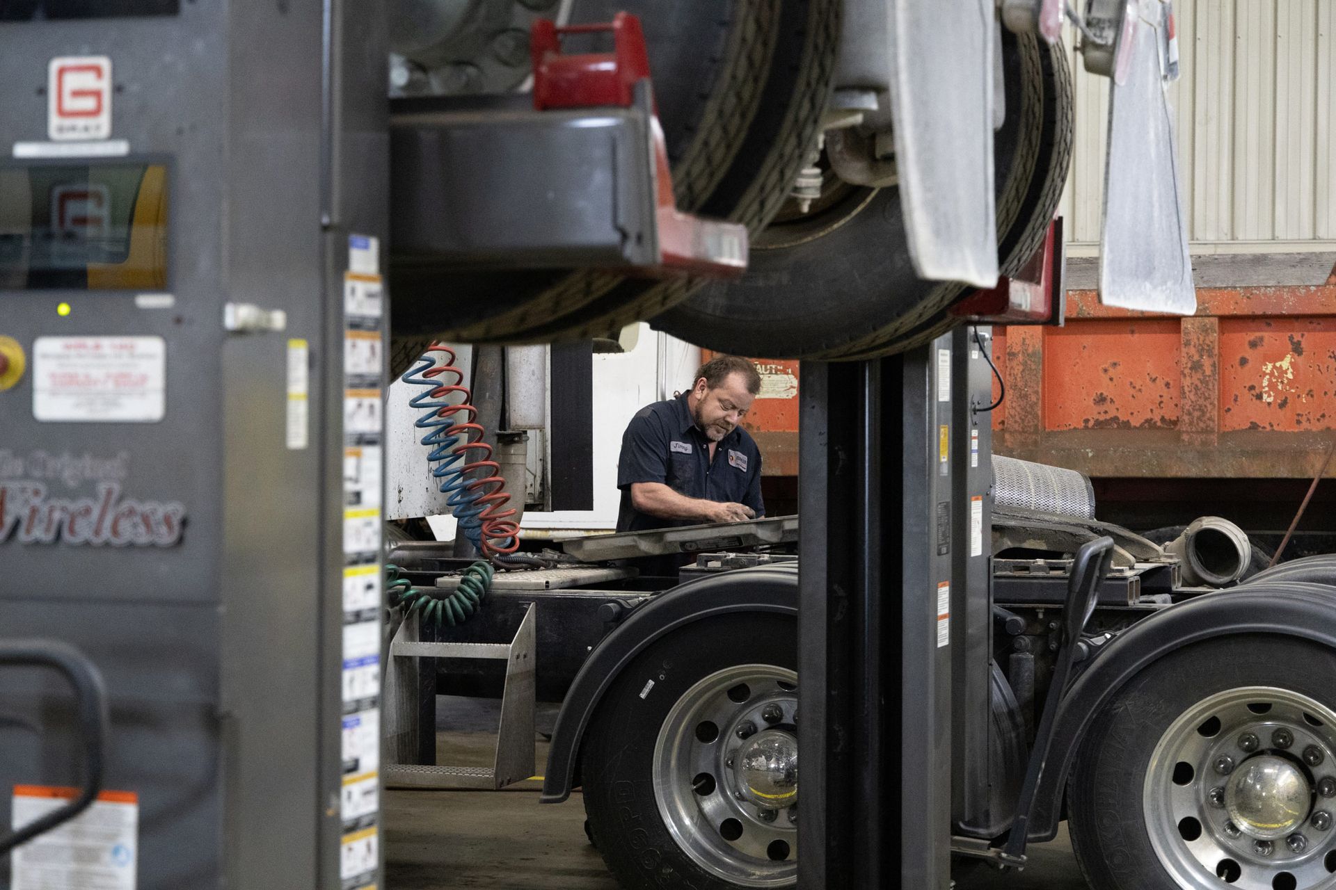 A mechanic working on a raised semi-truck in a repair shop. The mechanic is wearing dark clothing.