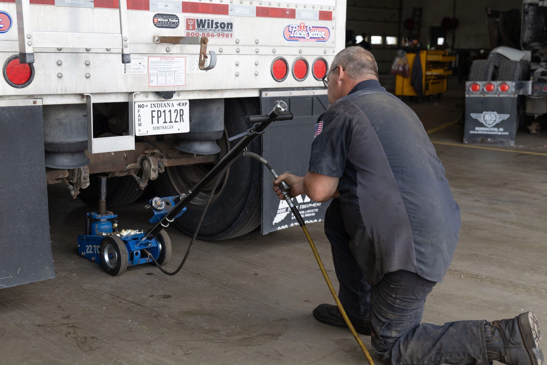 A mechanic kneels, working on the undercarriage of a truck. He is using a blue jack, with a black hose extending toward him.