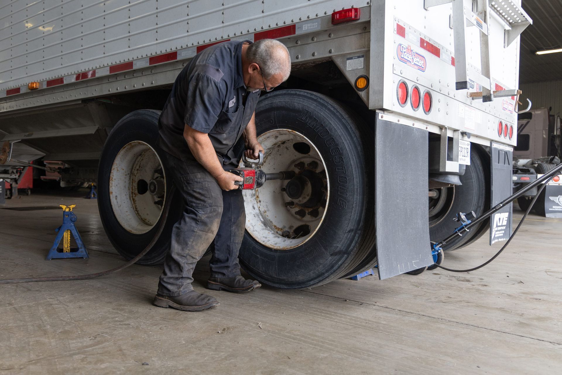 A mechanic uses a wrench to remove a tire from a semi-truck in a garage. He's wearing work clothes.
