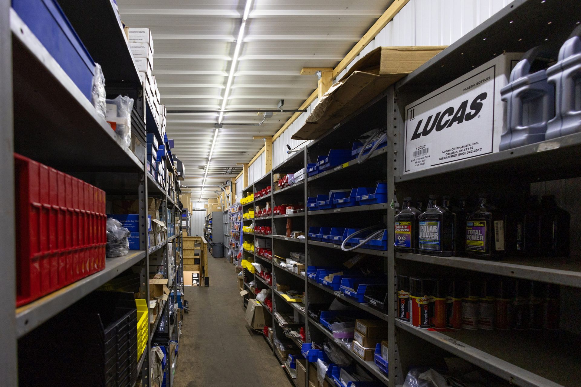 A narrow warehouse aisle filled with shelves stocked with various automotive parts and boxes, including some labeled 